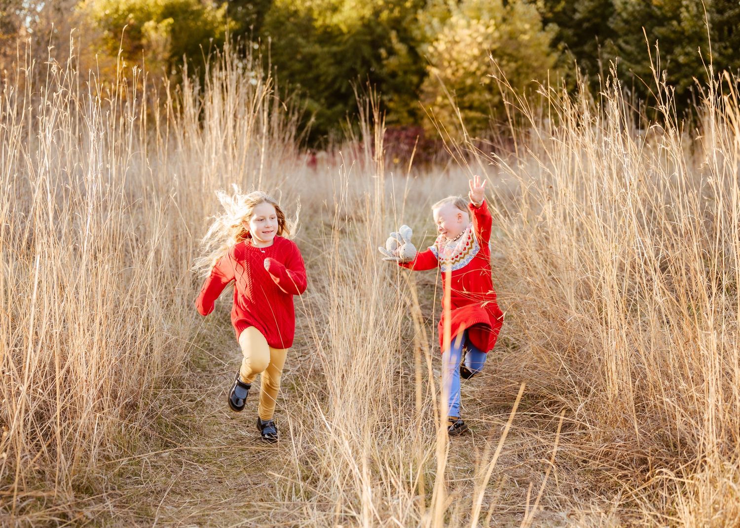 Two young girls in red coats running through tall golden grass on a sunny autumn day.