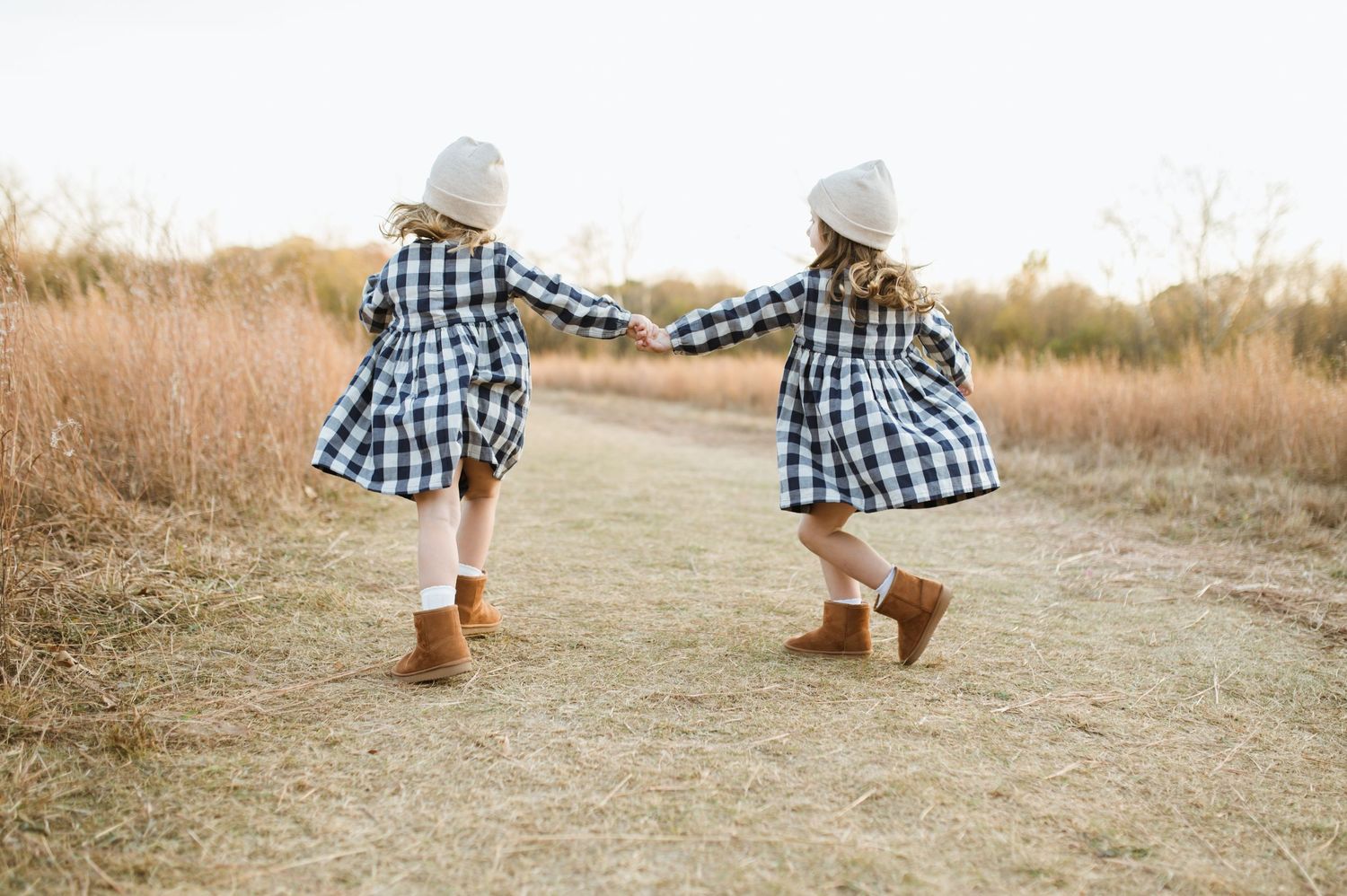 Two young girls in matching plaid dresses and white beanies holding hands on a fall field path.