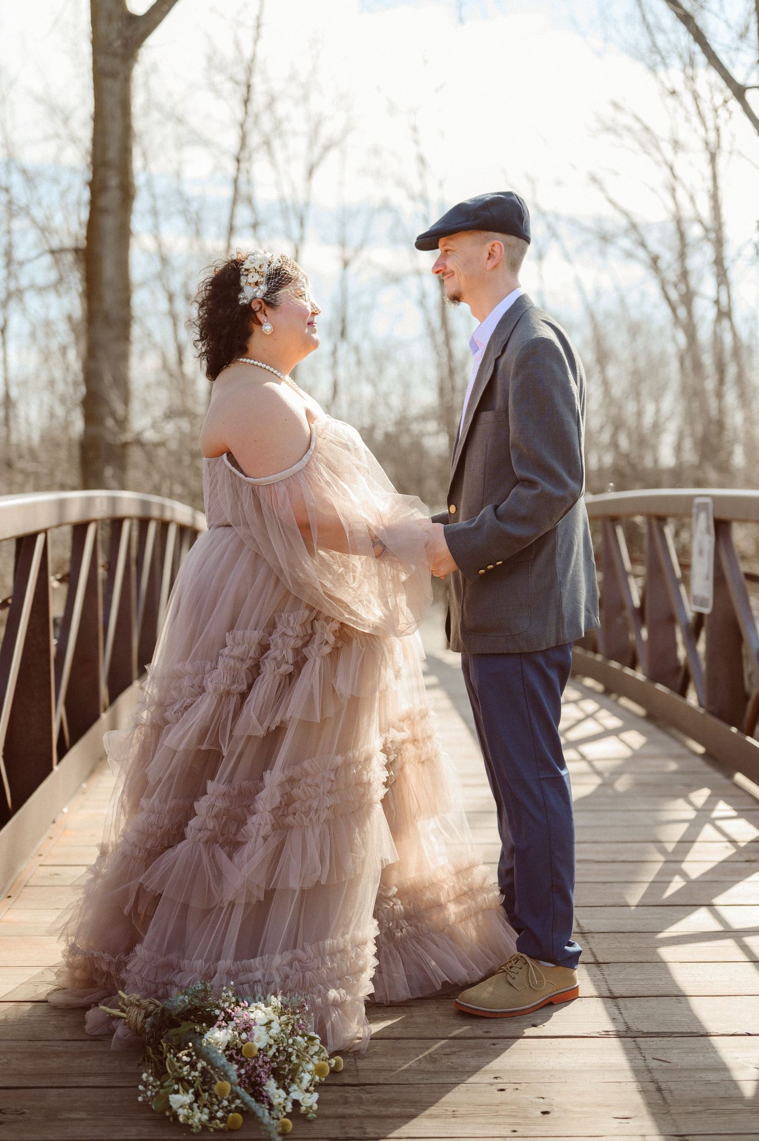 Bride in blush ruffled tulle gown holding hands with groom on a wooden bridge in winter.