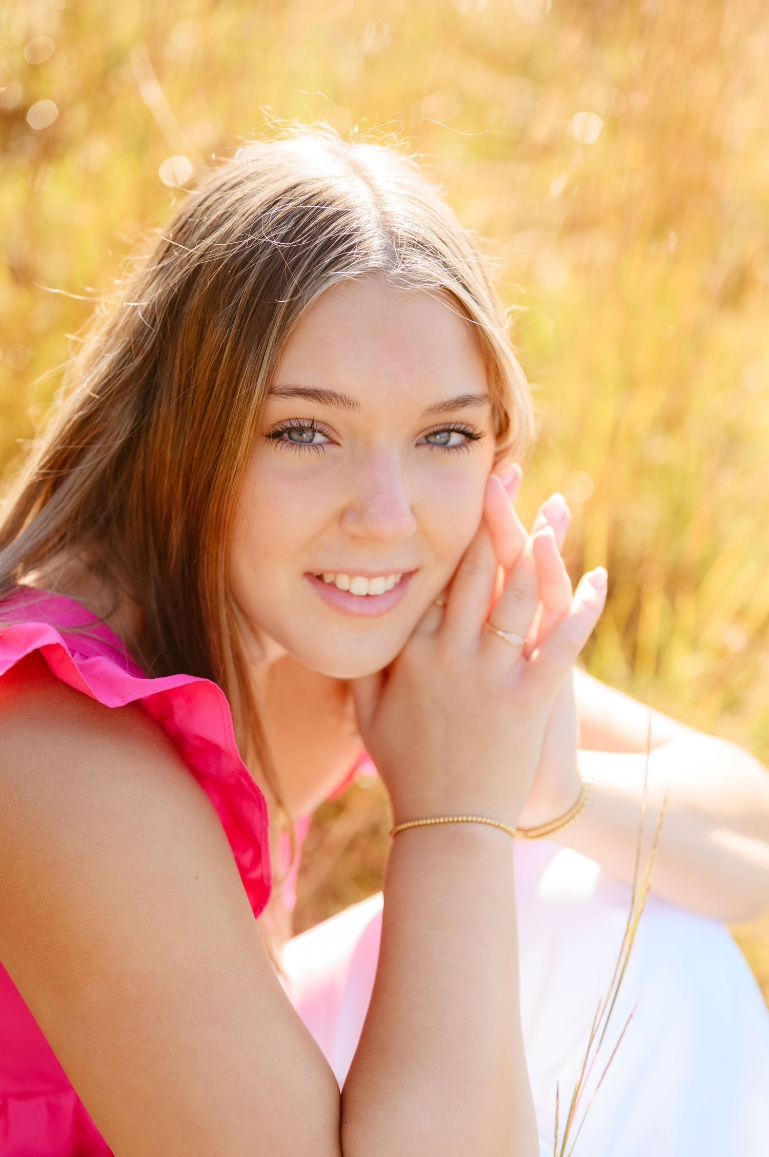 Smiling teen girl in pink top with gold bracelet poses outdoors in warm golden sunlight.