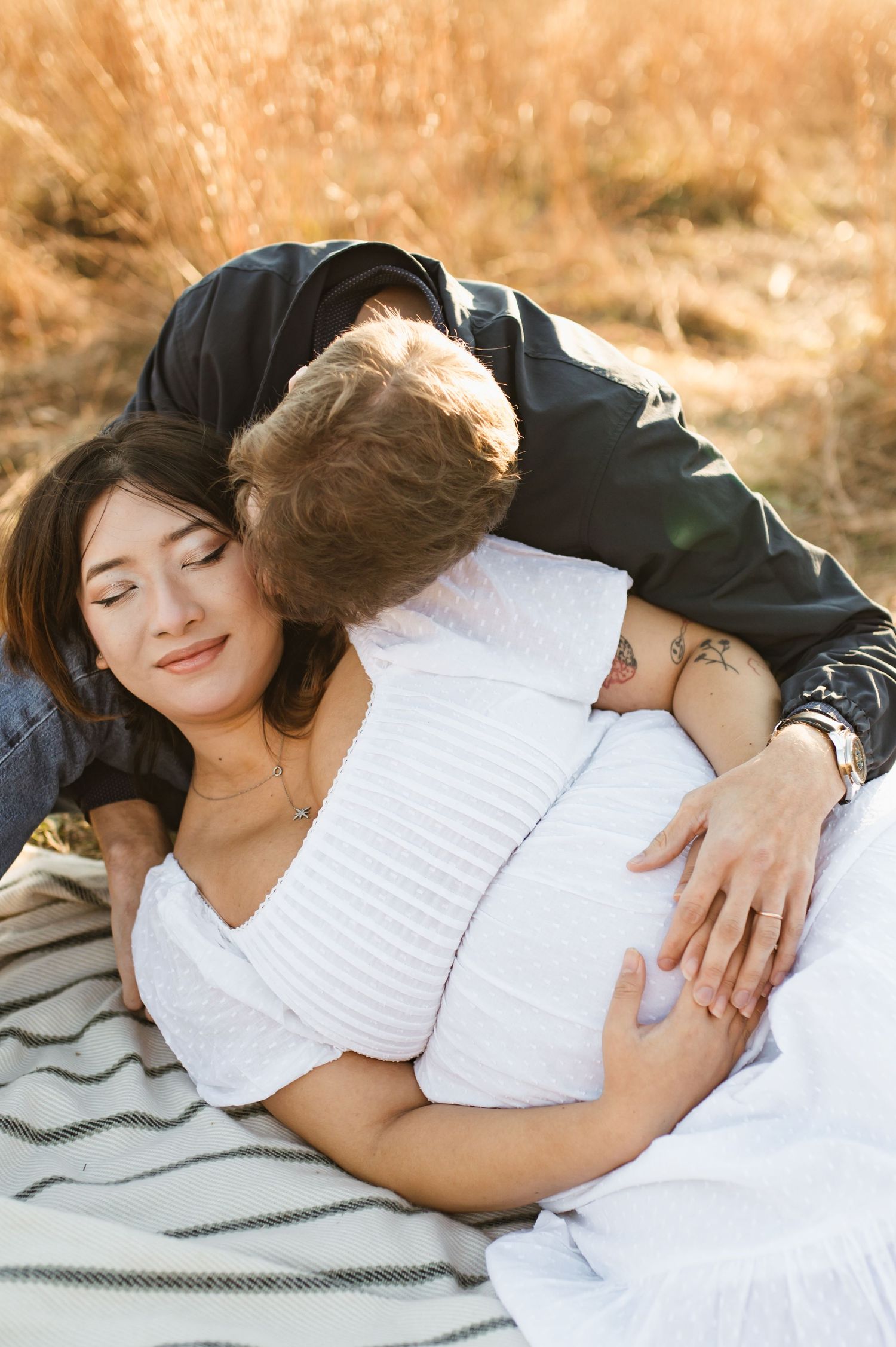 A man tenderly kisses a pregnant woman as she reclines on a blanket in a golden field.