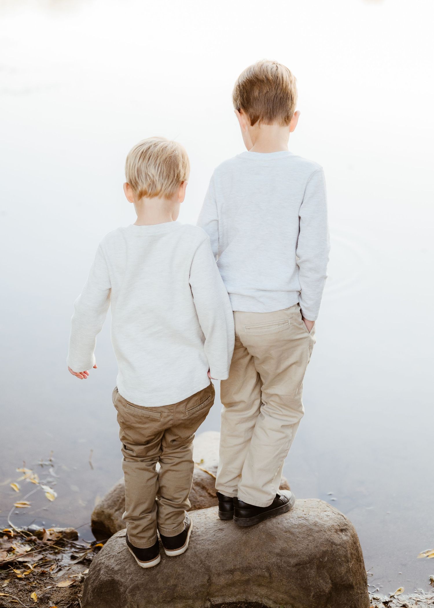 Two young boys in matching outfits standing on a rock gazing at a calm misty lake together.
