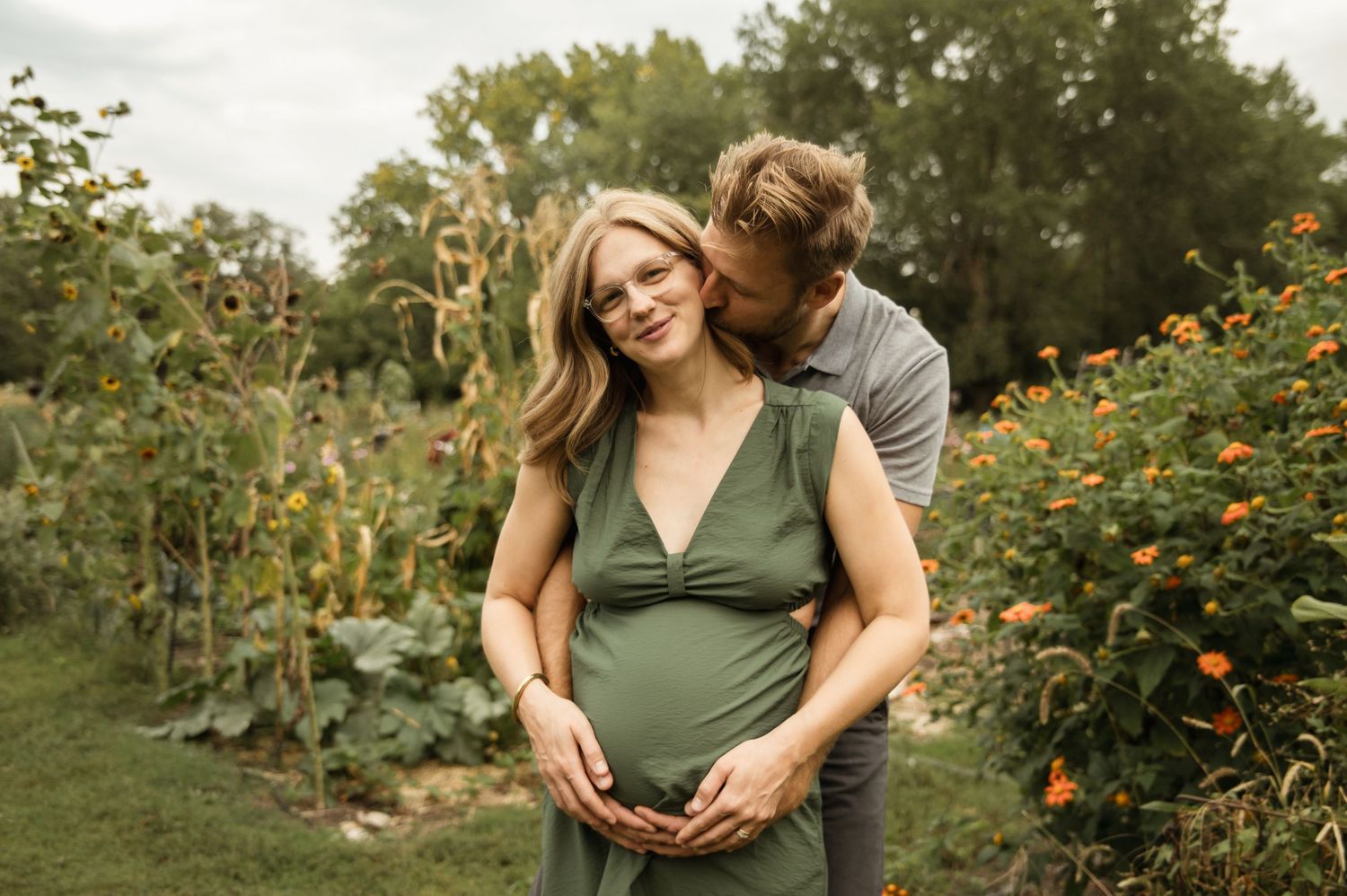 Pregnant woman in green dress embraced by partner in lush garden with orange flowers during maternity photoshoot.