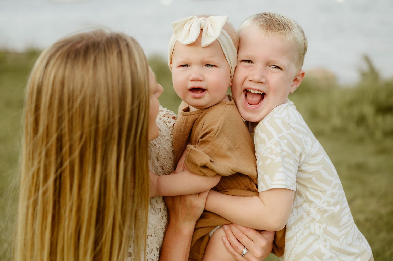 Laughing mother hugs her two young children, a baby girl and toddler boy, outdoors near water. Minneapolis Family Photographer
