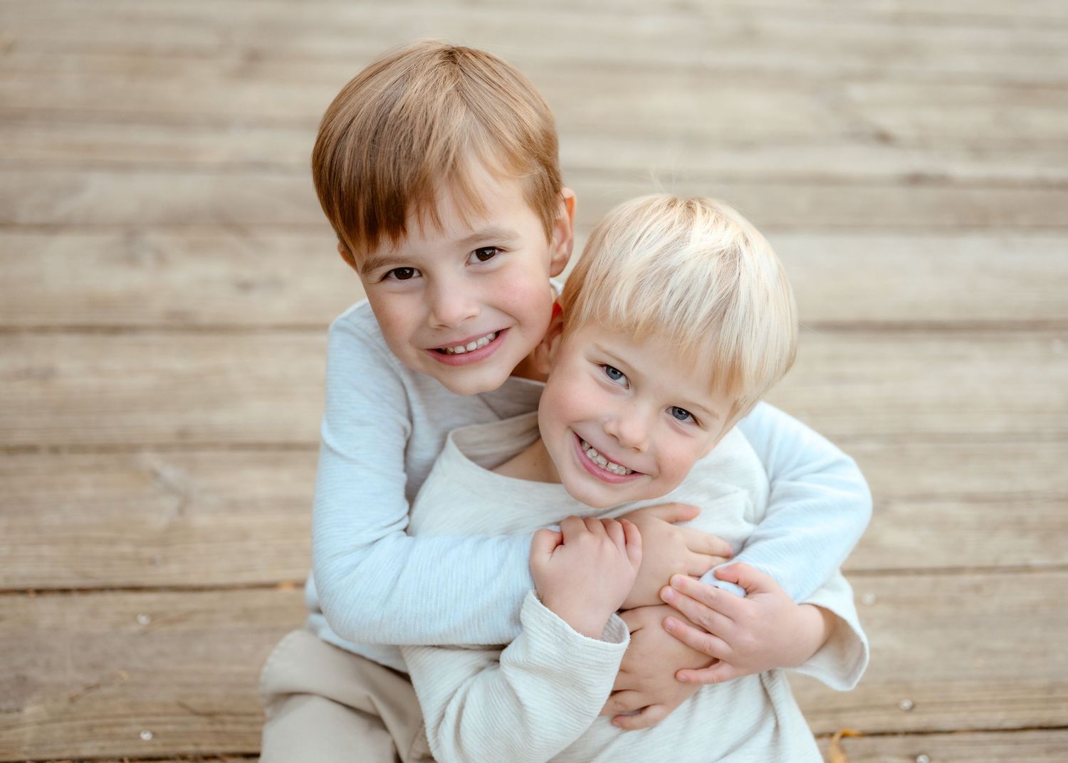 Two young red and blonde-haired brothers hugging and smiling on a wooden deck in matching outfits.