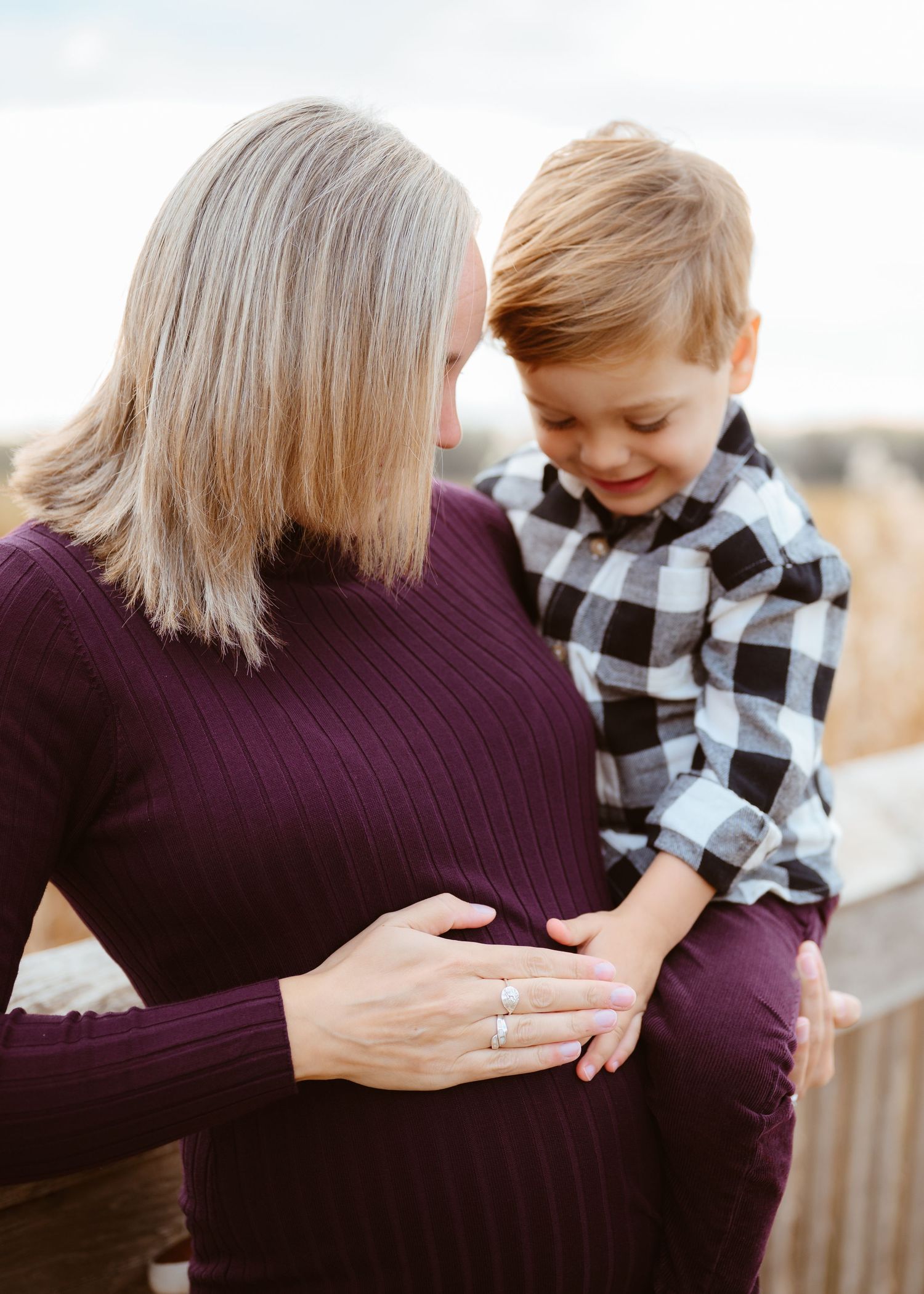 Pregnant woman in burgundy dress holds toddler boy who touches her baby bump during outdoor maternity photo session.
