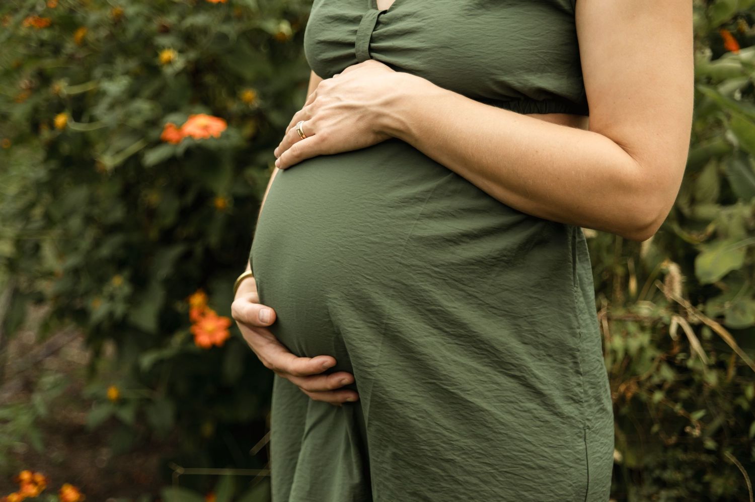 Pregnant woman in an olive green dress gently holding her baby bump in a garden setting.