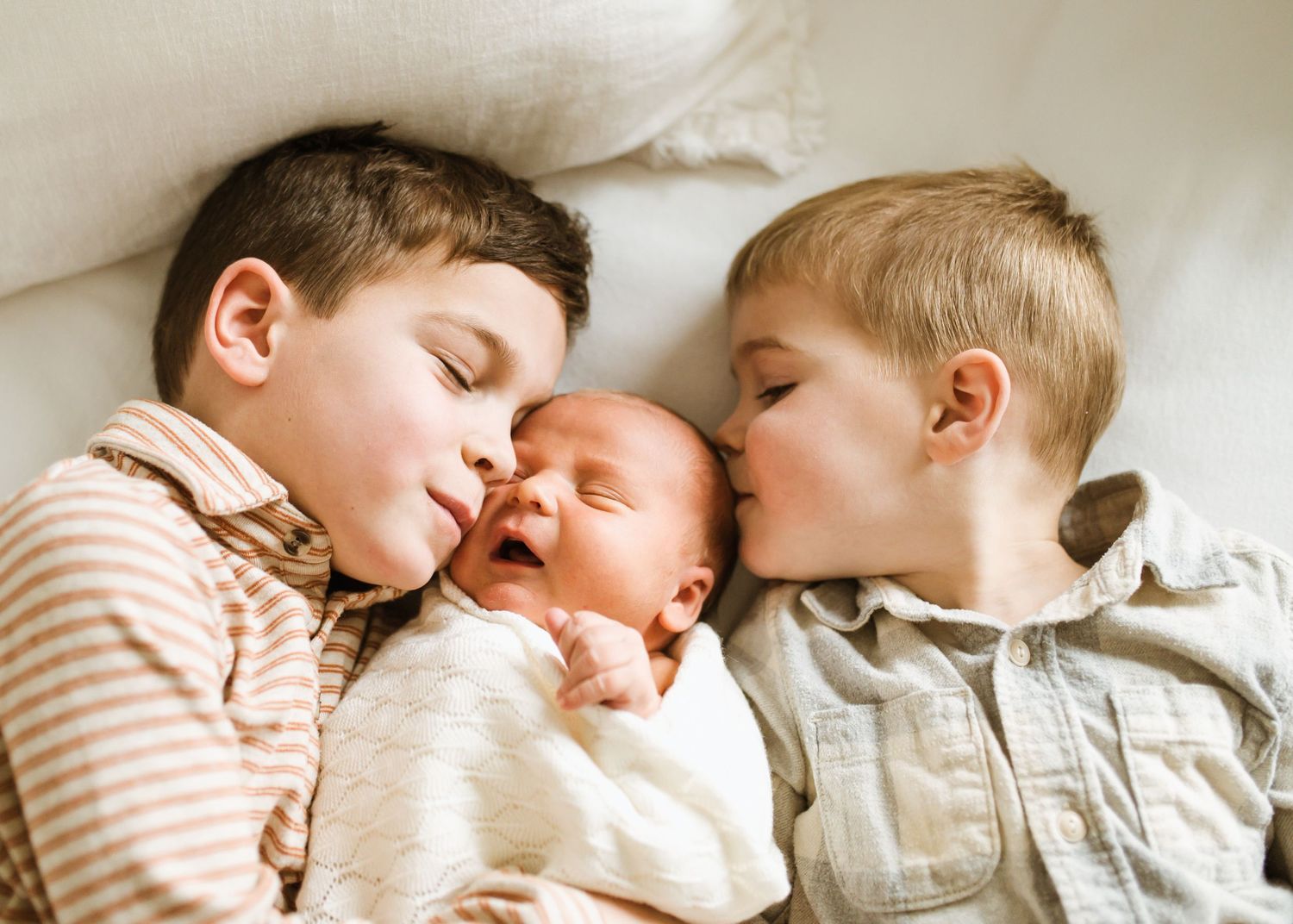 Two brothers lying beside laughing swaddled newborn baby on white bedding.