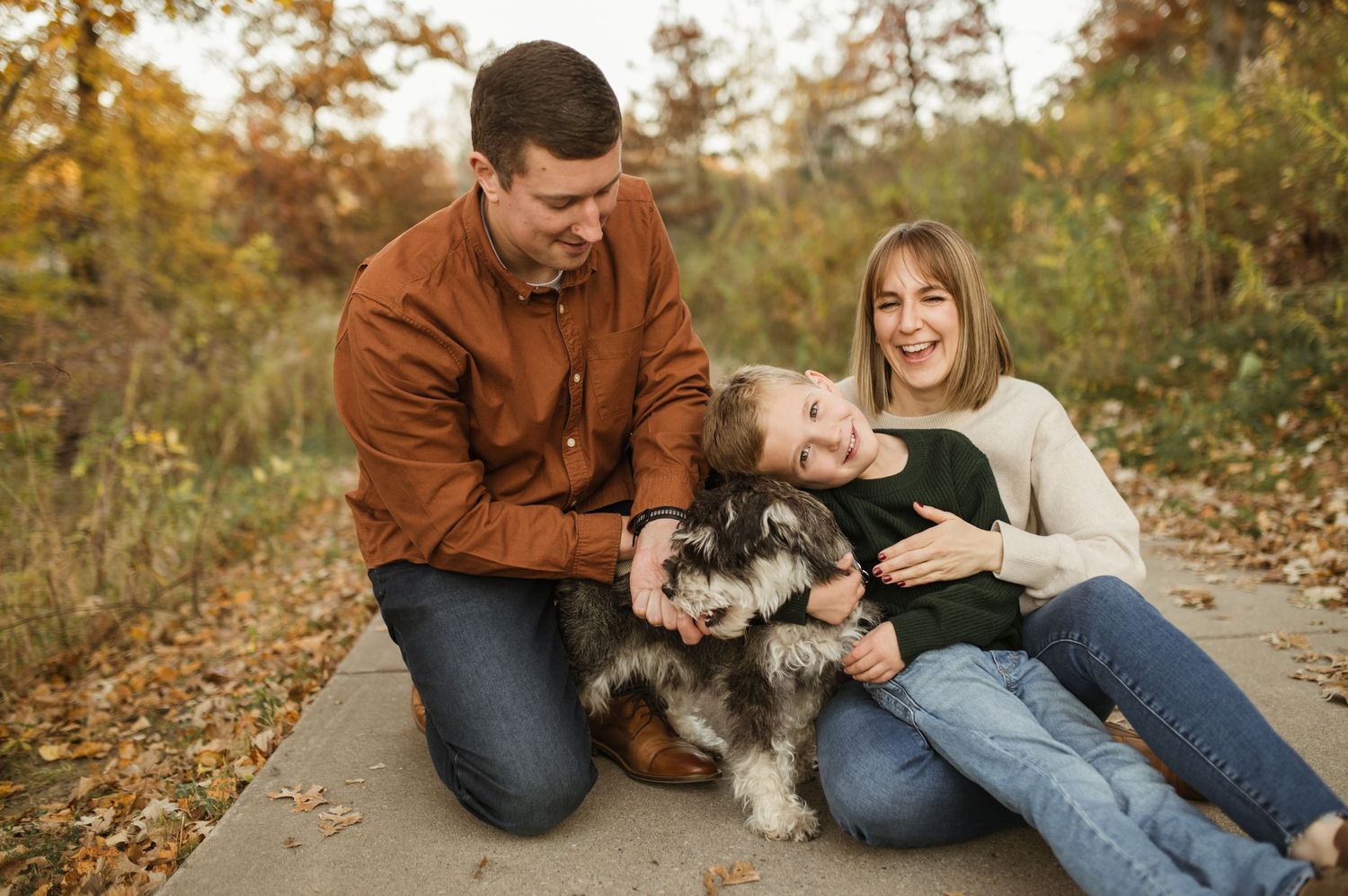 A happy family with a fluffy dog sits together on a path surrounded by autumn foliage.