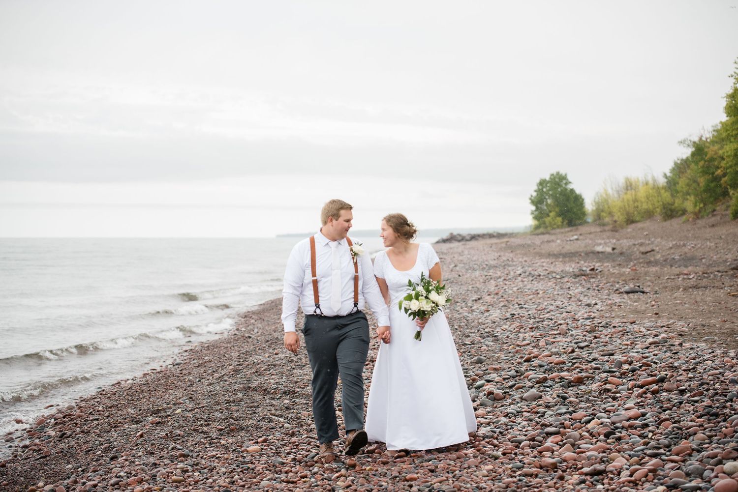 Newlywed couple walks hand-in-hand along rocky shoreline, bride holding bouquet, groom wearing suspenders.