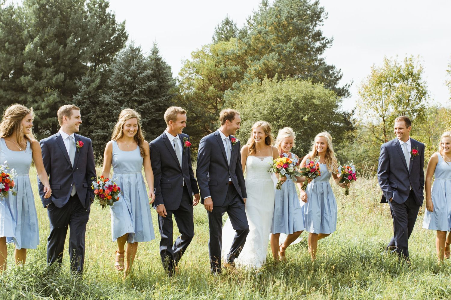 Wedding party walking through green field with bridesmaids in light blue dresses and groomsmen in navy suits.