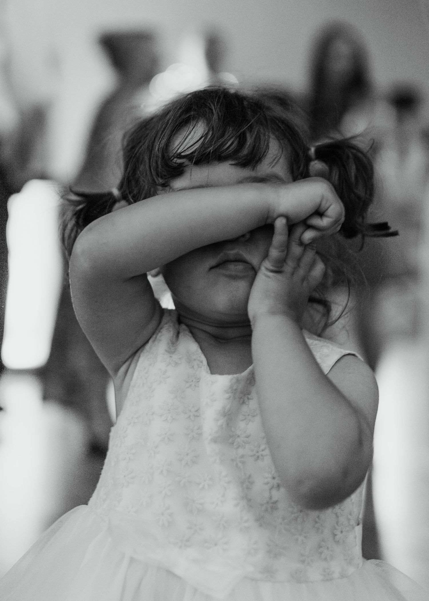 Adorable flower girl in white dress covering her face with her arms at wedding reception.