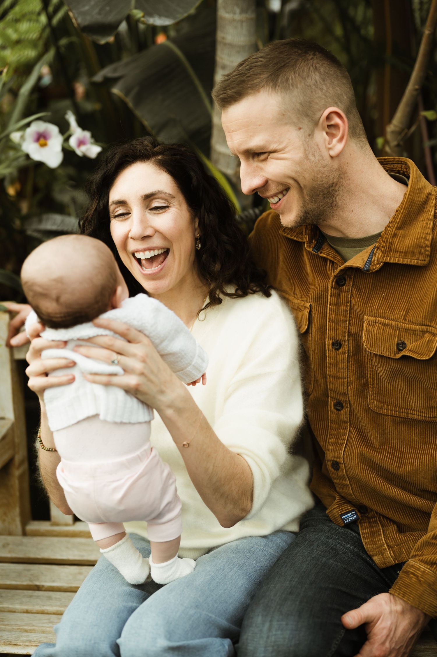 Laughing parents sitting outdoors holding newborn baby surrounded by tropical plants.