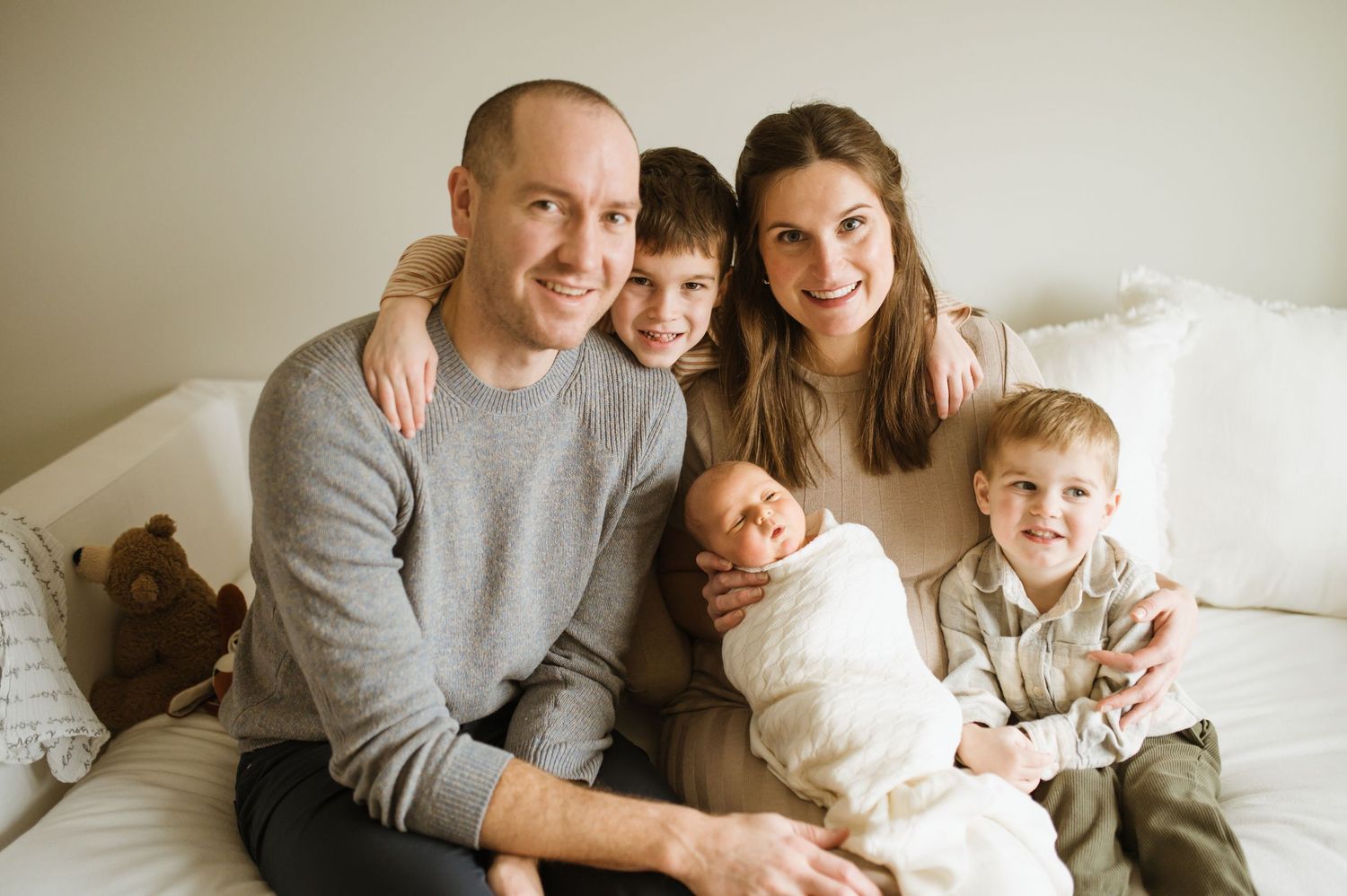 Happy family of five with newborn baby sitting together on white bed.