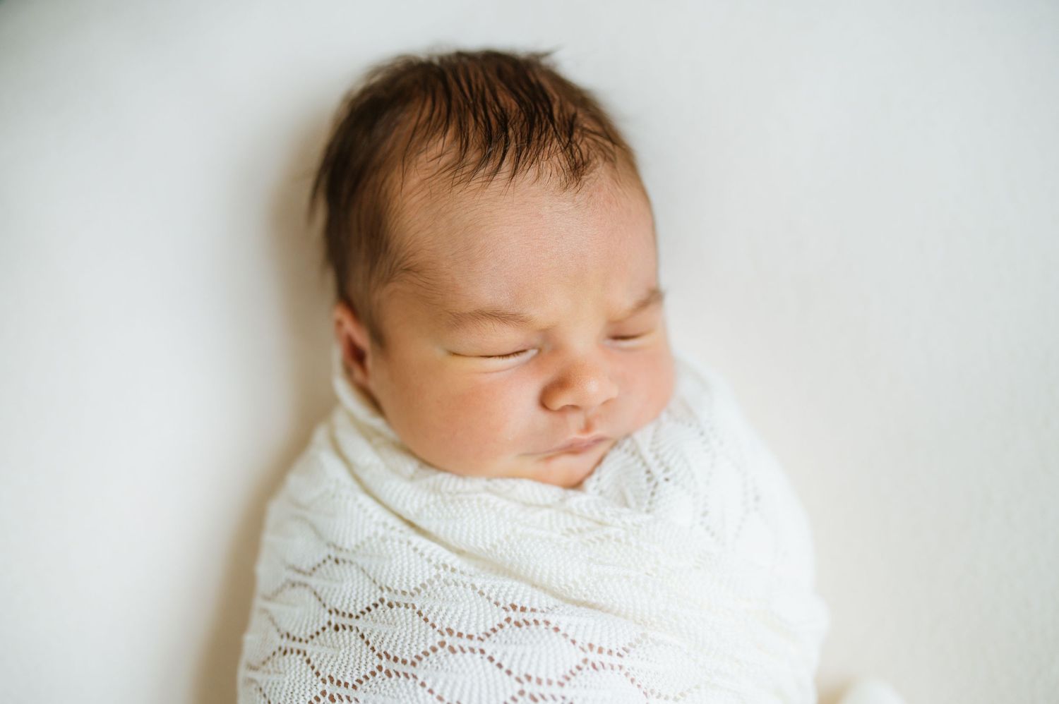 A peaceful newborn baby sleeps swaddled in a soft white knit blanket.