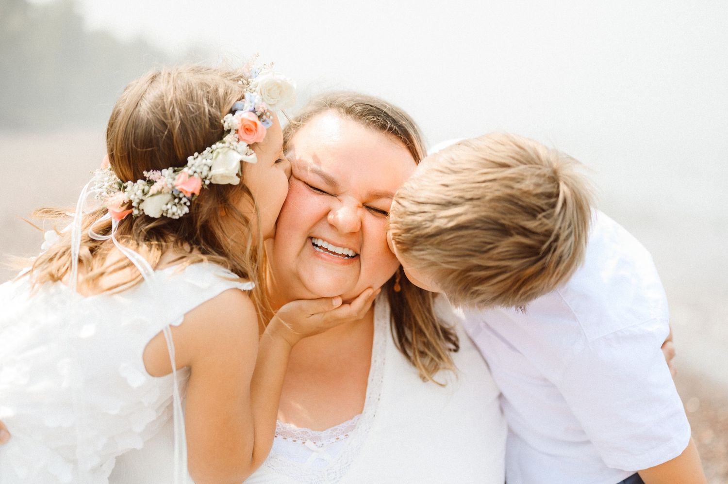 Two children kiss a laughing mother on both cheeks simultaneously in a joyful outdoor portrait.