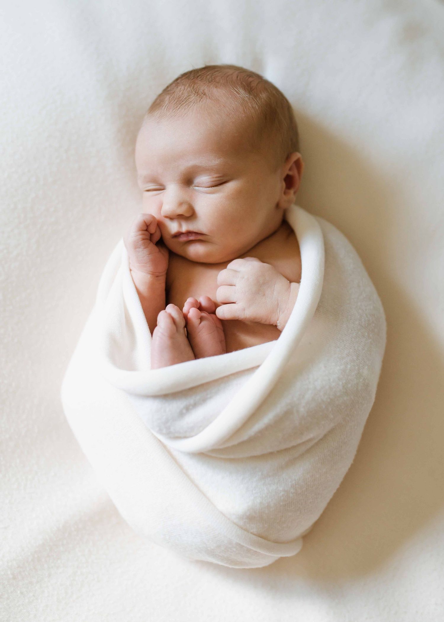 Newborn baby swaddled in a white wrap, sleeping peacefully on a soft white blanket.