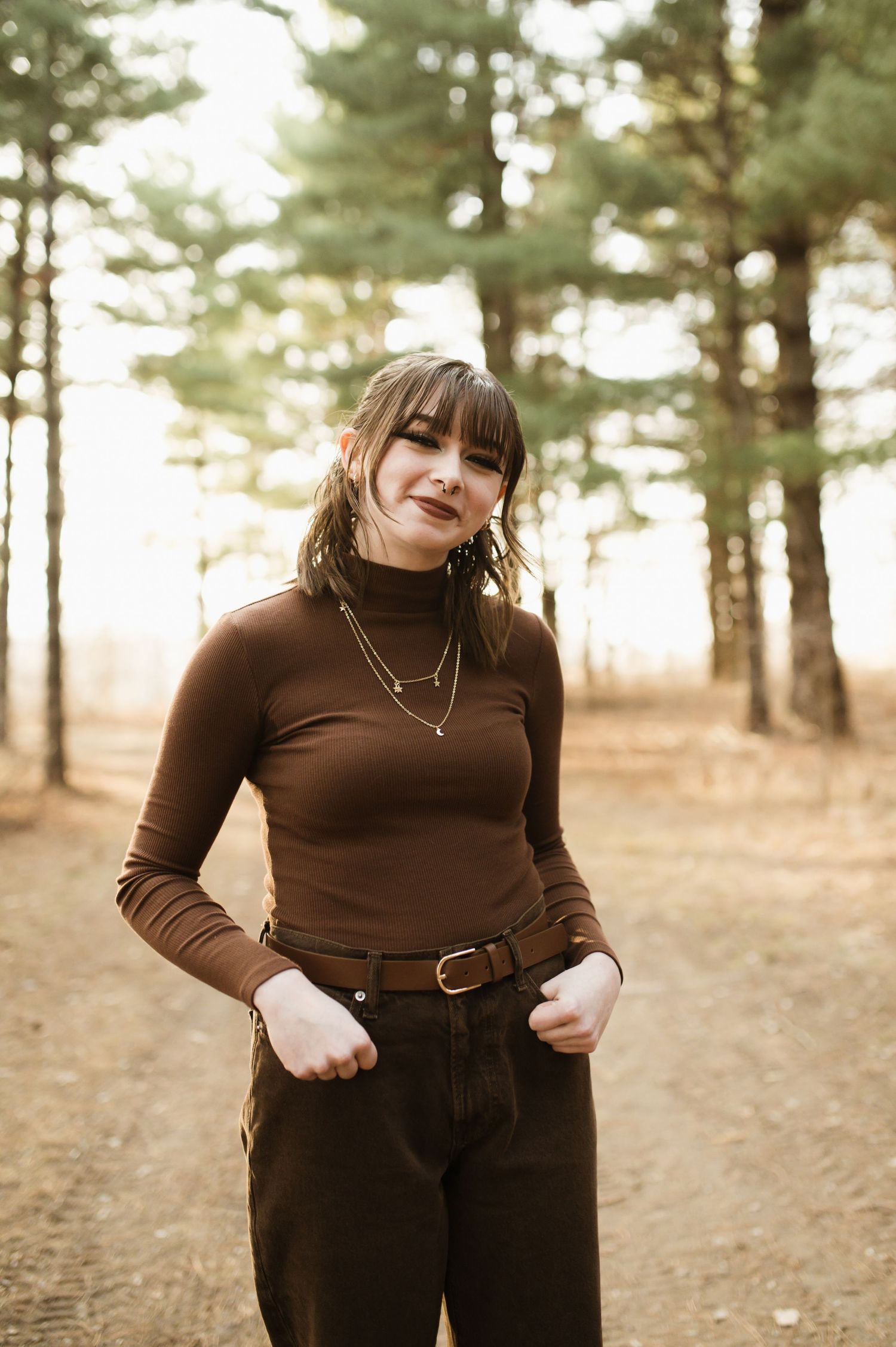 Young woman in brown turtleneck and dark pants poses confidently on forest path among tall pine trees.