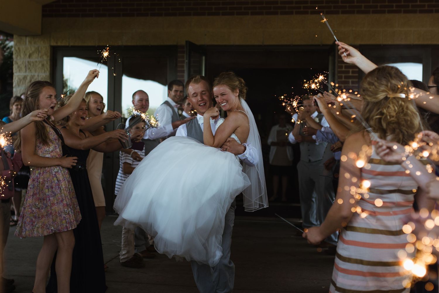Groom carrying bride through sparkler send-off surrounded by cheering wedding guests at night.