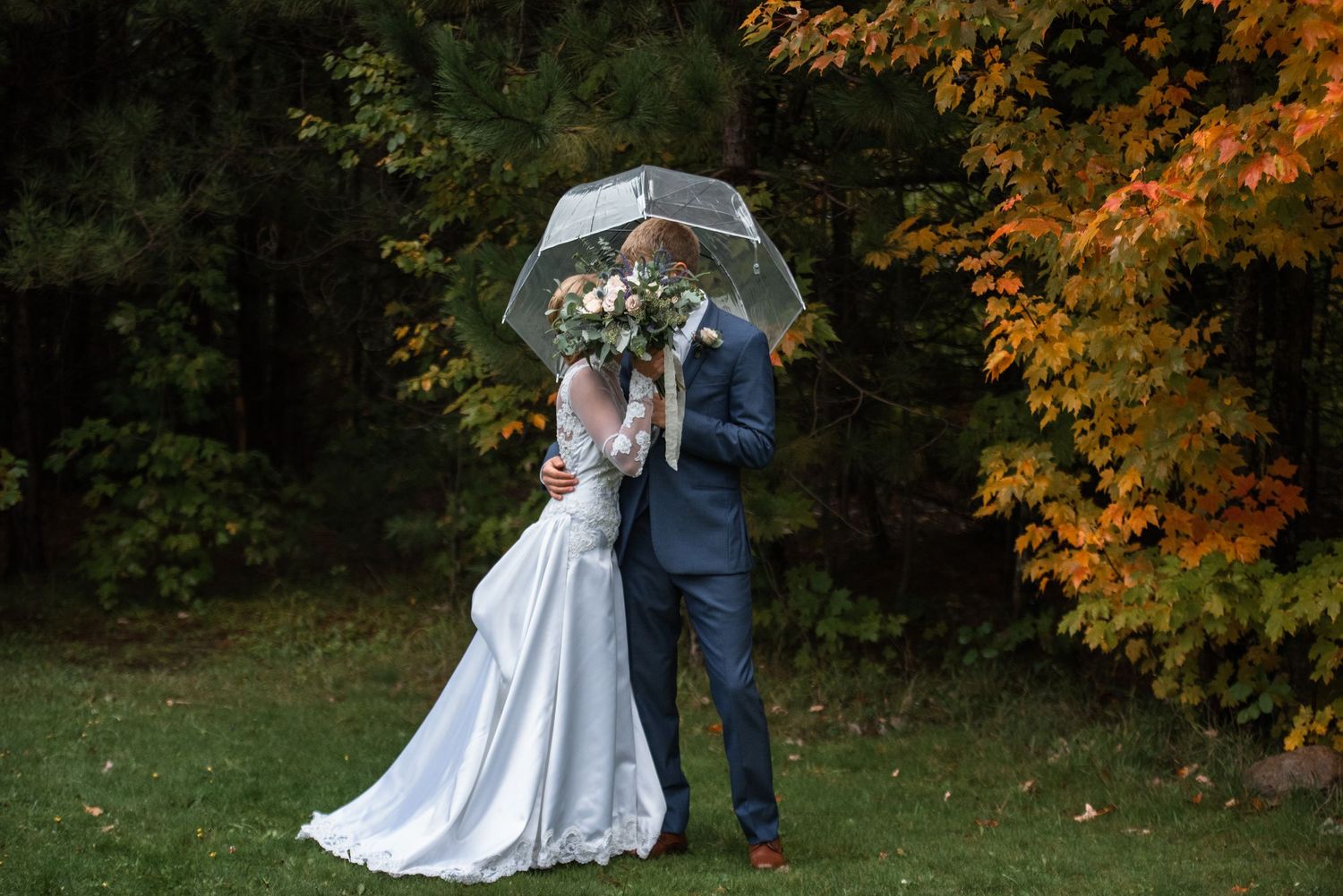 Bride and groom share a kiss under a clear umbrella in autumn garden with colorful fall foliage.
