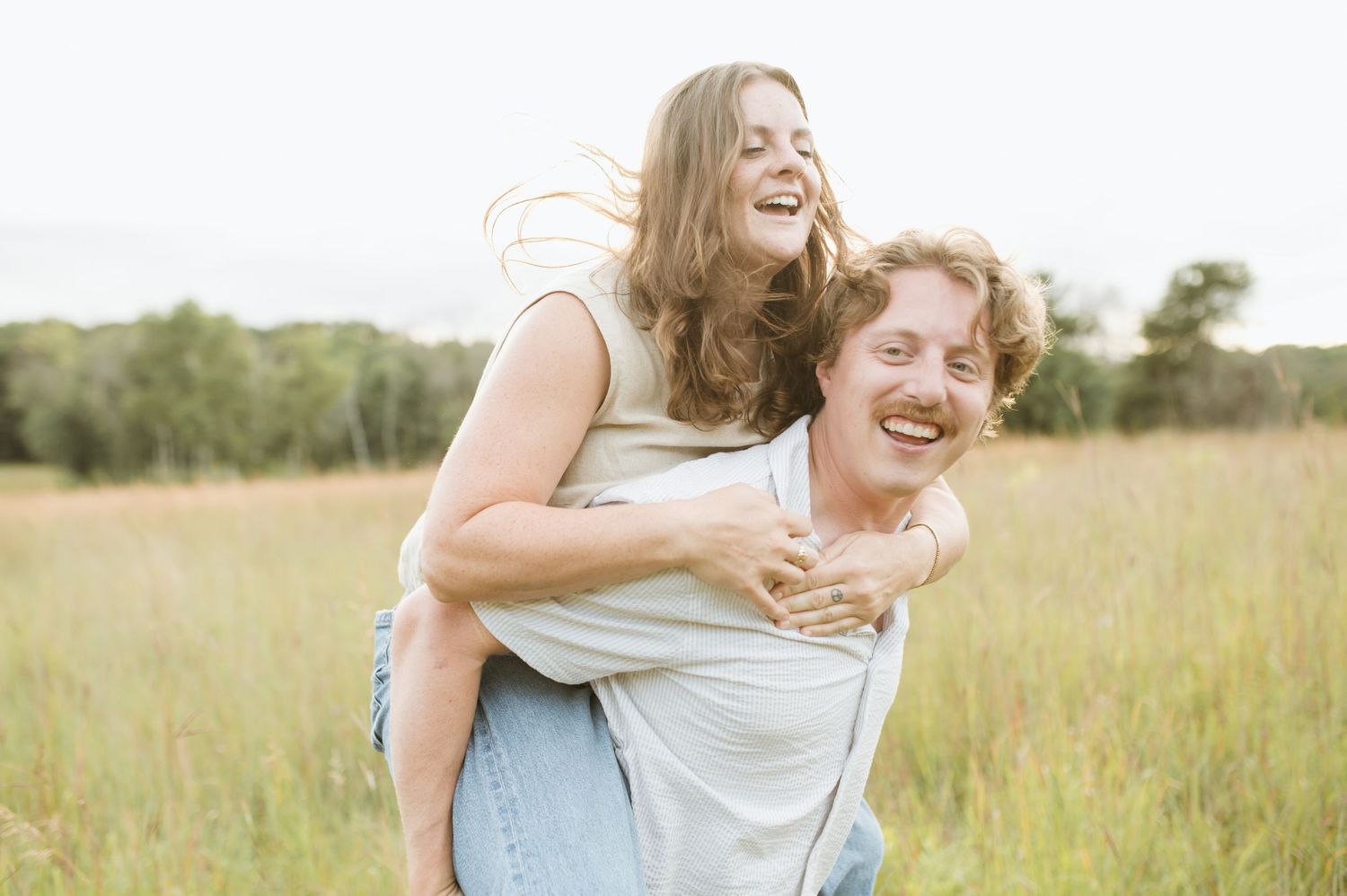 A joyful couple laughs together as the man carries the woman on his back in a sunny meadow.