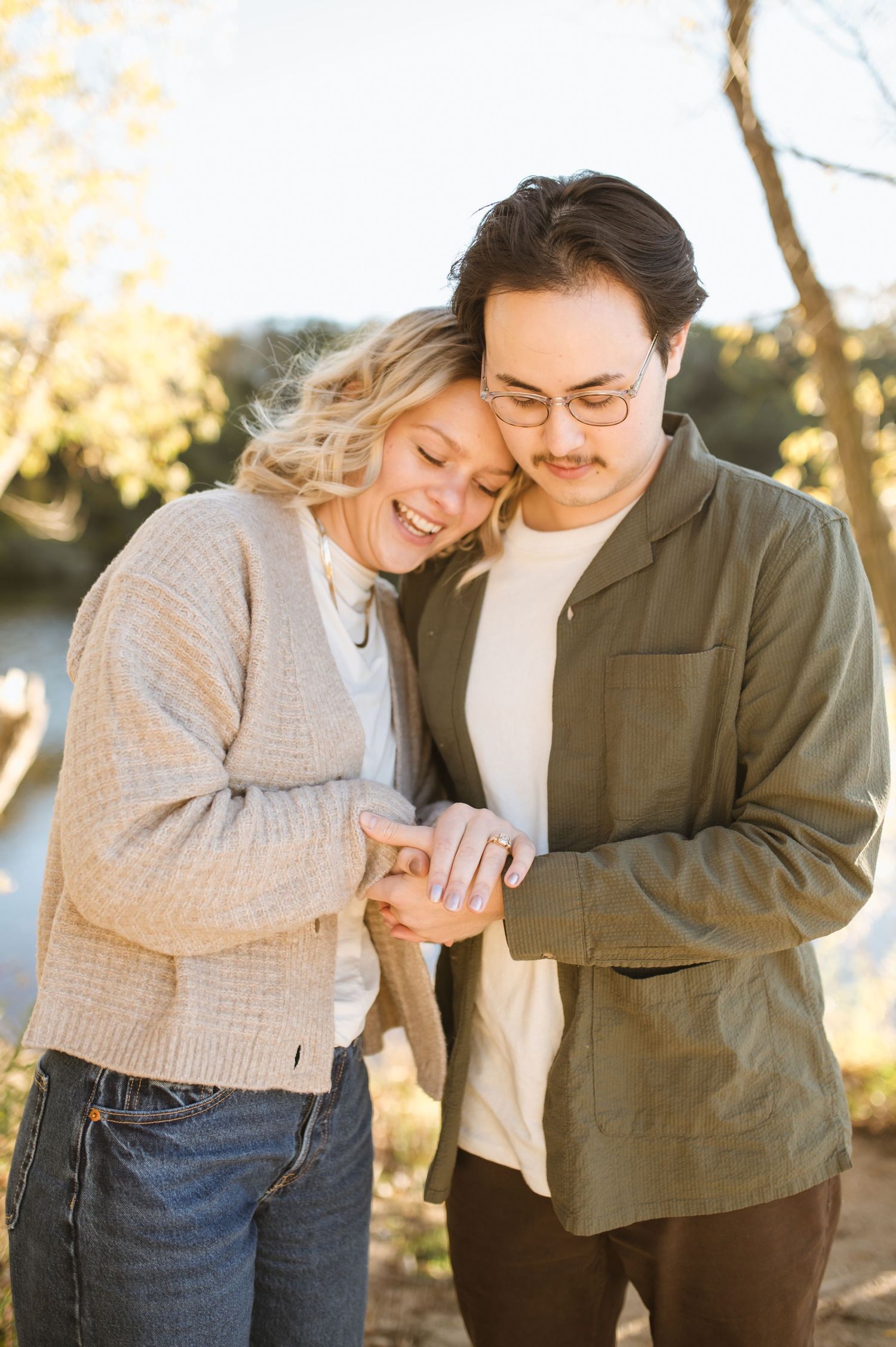 A happy couple looks down at an engagement ring while embracing outdoors in warm golden sunlight.