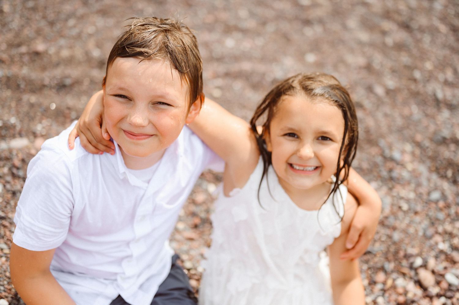 A boy and girl in white outfits smile together sitting on a pebbly ground outdoors.