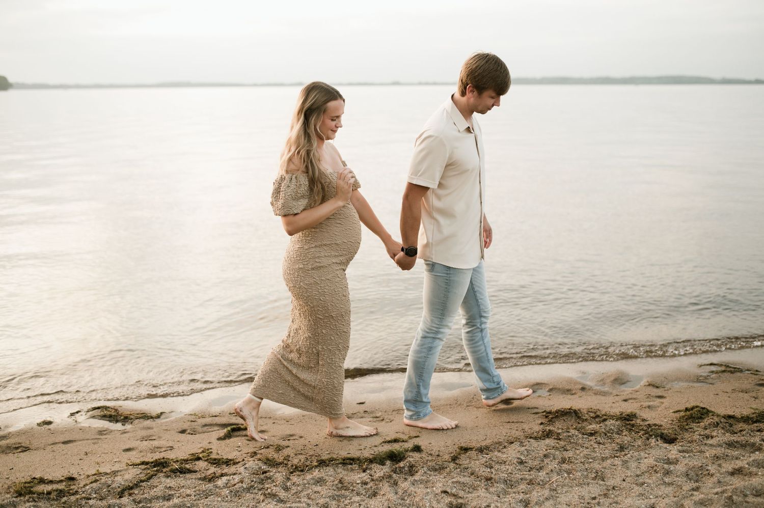 Pregnant couple holding hands walking barefoot along a sandy beach at sunset.