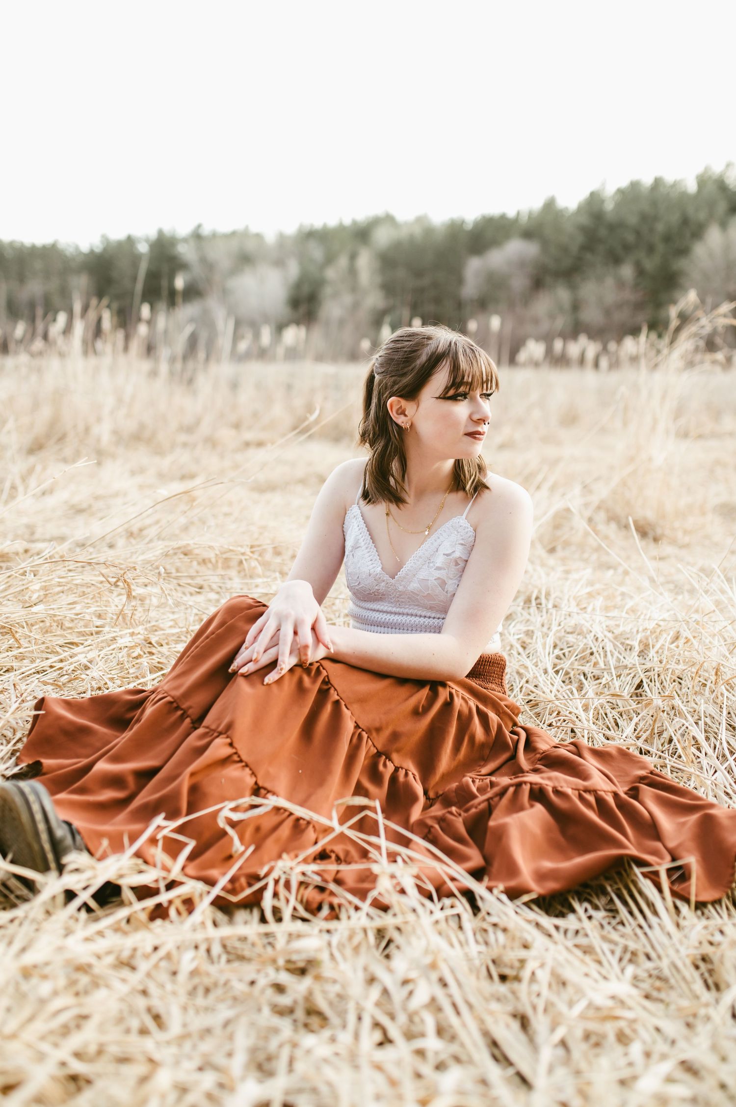 Young woman in rust maxi skirt and white lace top sits relaxed in golden hay field outdoors.