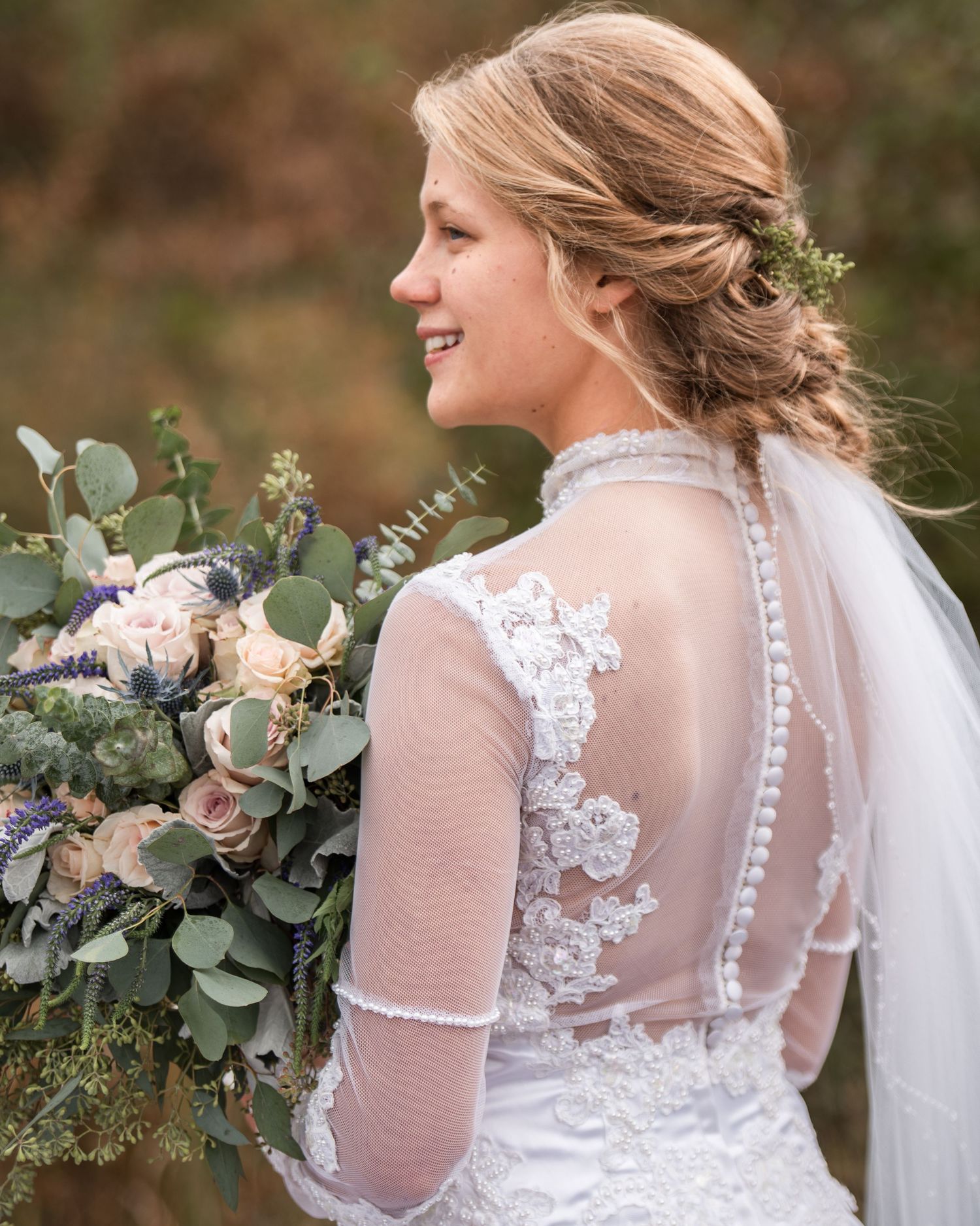 Bride in lace long-sleeve wedding gown with button back detail holds lush greenery and blush bouquet.