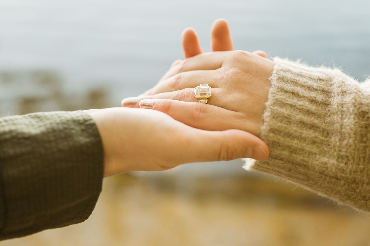 A man gently holds a woman's hand displaying a gold emerald-cut engagement ring outdoors by water.