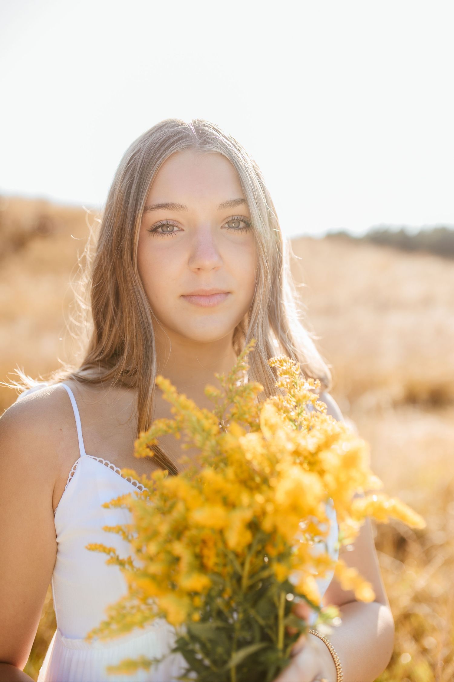 Teen girl with long blonde hair holds bouquet of yellow wildflowers in sunlit golden field, gazing at camera.