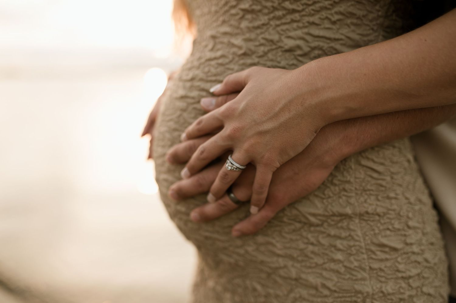 Pregnant woman in beige dress with partner's hands cradling baby bump, wearing engagement ring.