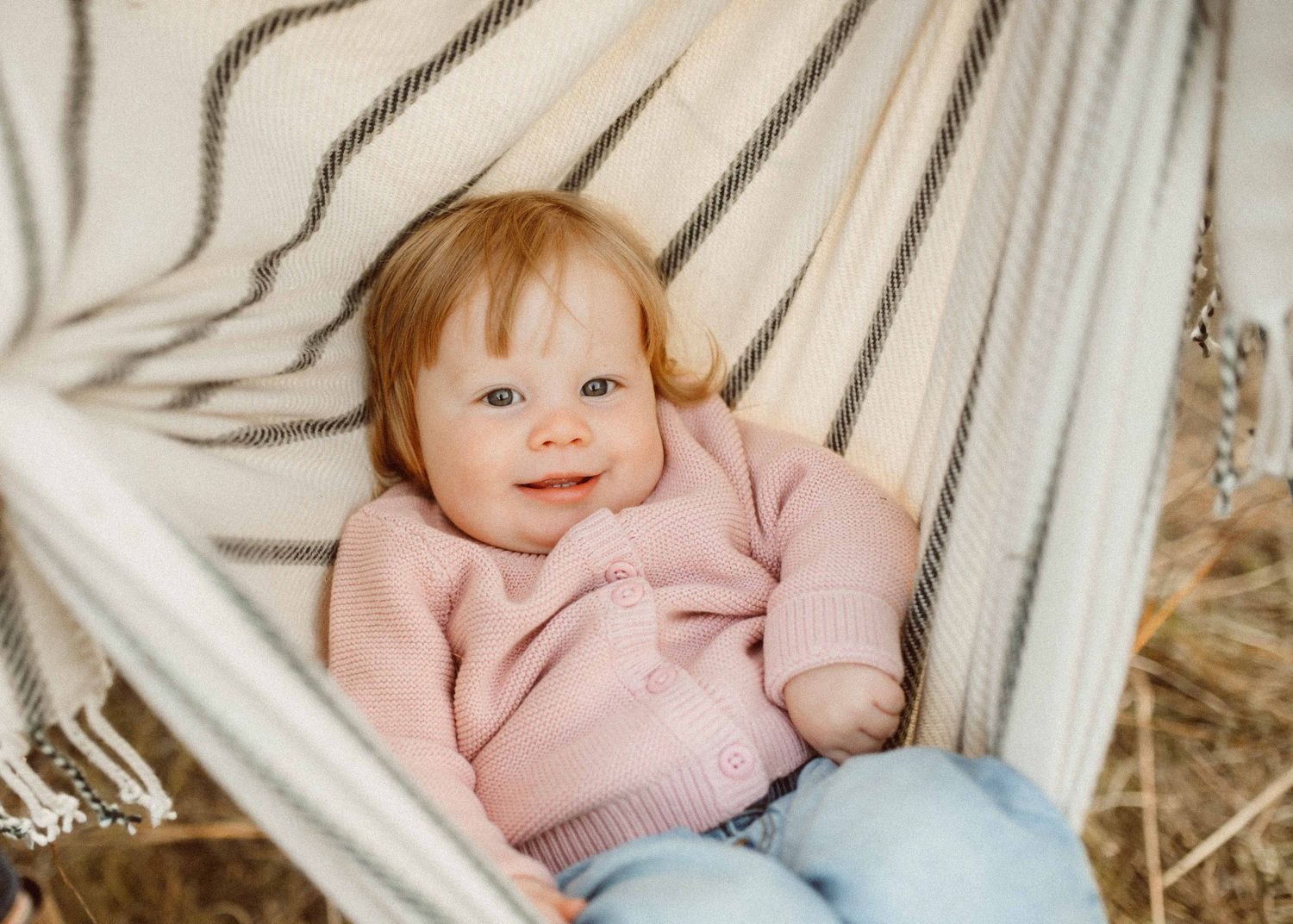 Baby girl in pink sweater smiling in a striped hammock outdoors. Minneapolis Family Photography.