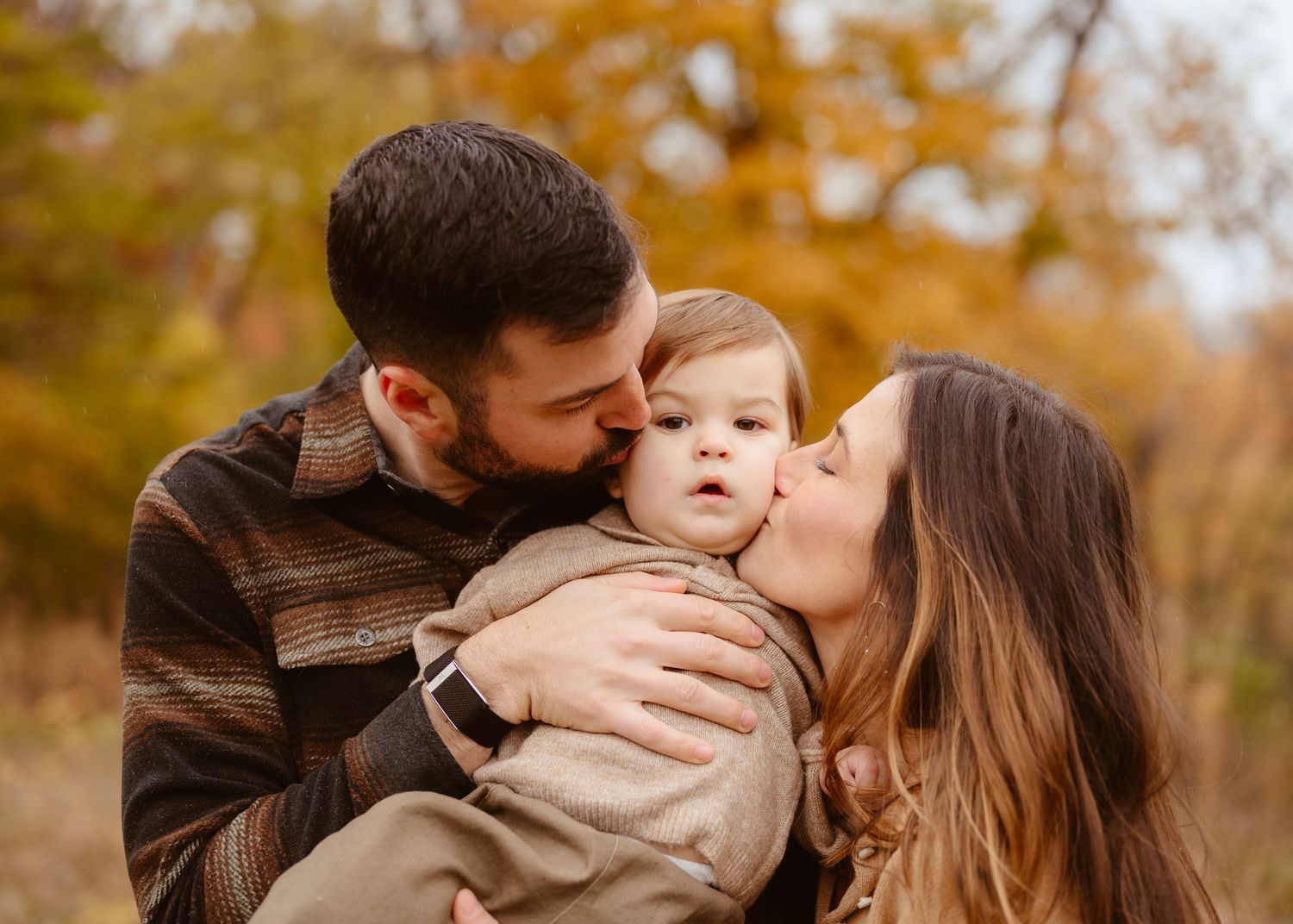 Young parents kissing their baby boy on the cheeks outdoors in a colorful autumn setting.