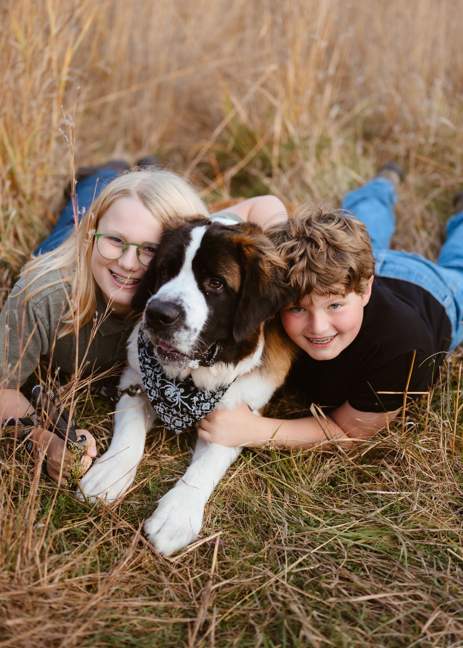 Two smiling children lying in golden grass with a Saint Bernard dog wearing a bandana outdoors.