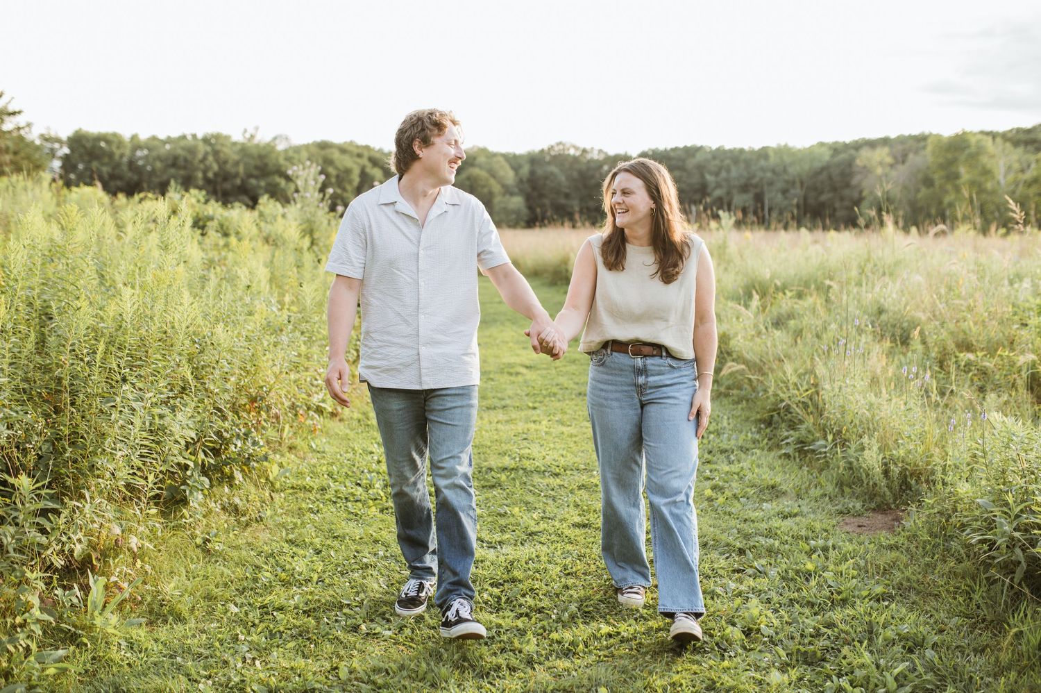 A young couple holds hands while walking along a grassy path through an open meadow together.