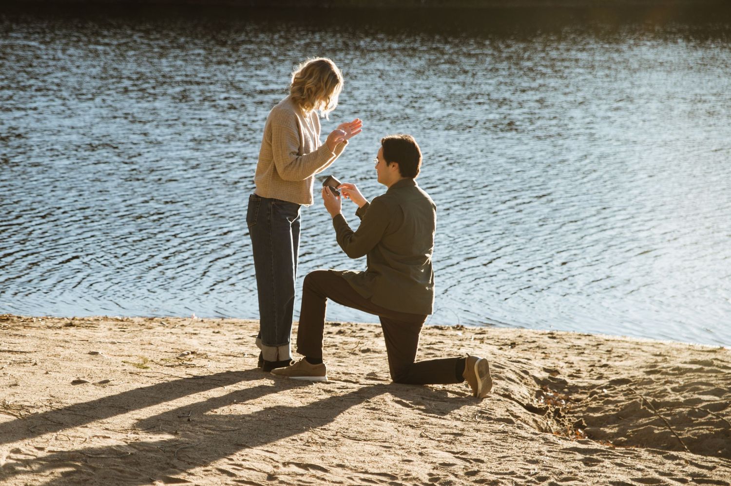 Man kneeling on one knee proposing to a woman on a sandy riverbank at golden hour.