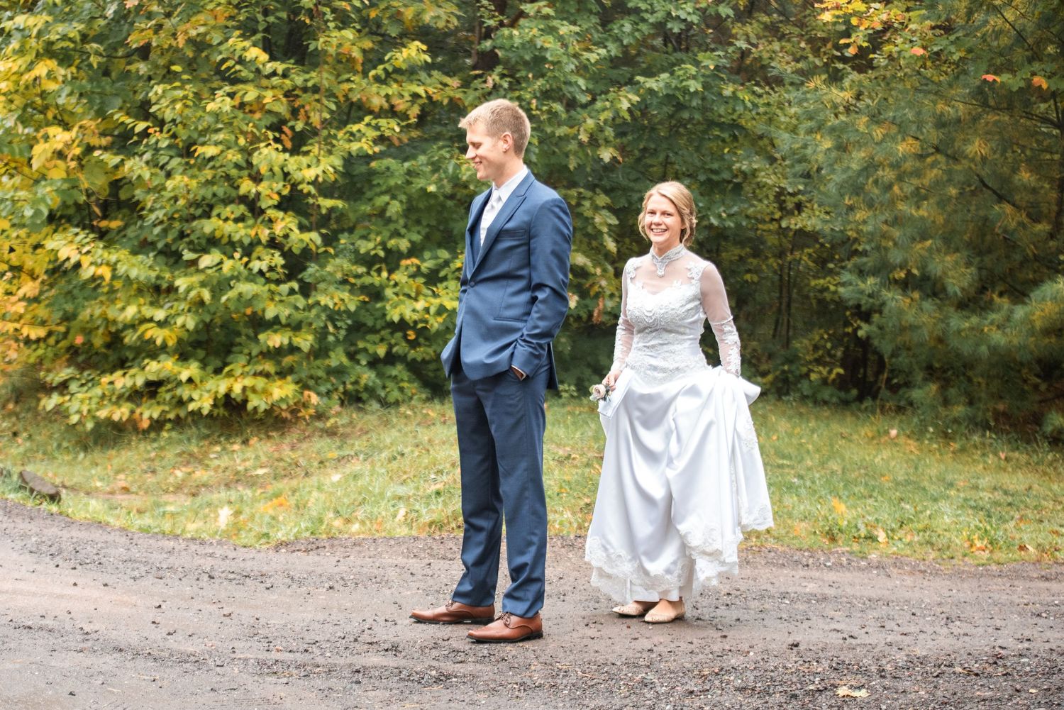 Bride in white gown and groom in blue suit walk together on gravel path with autumn foliage backdrop.