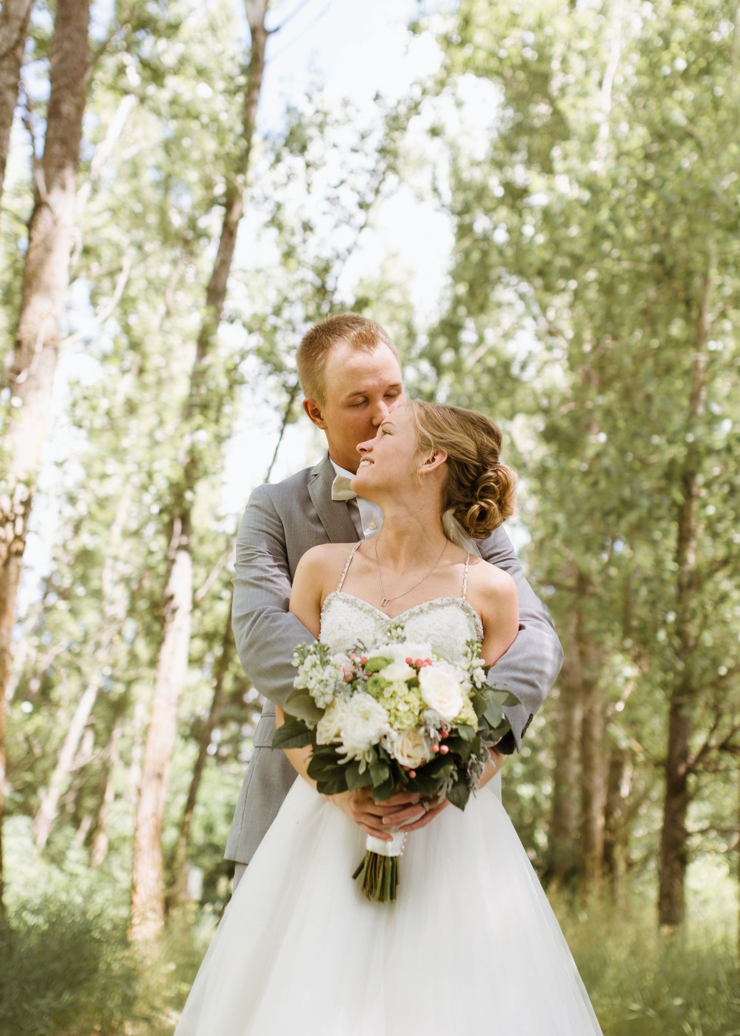 Groom in gray suit embracing bride in white gown while kissing in an aspen forest.