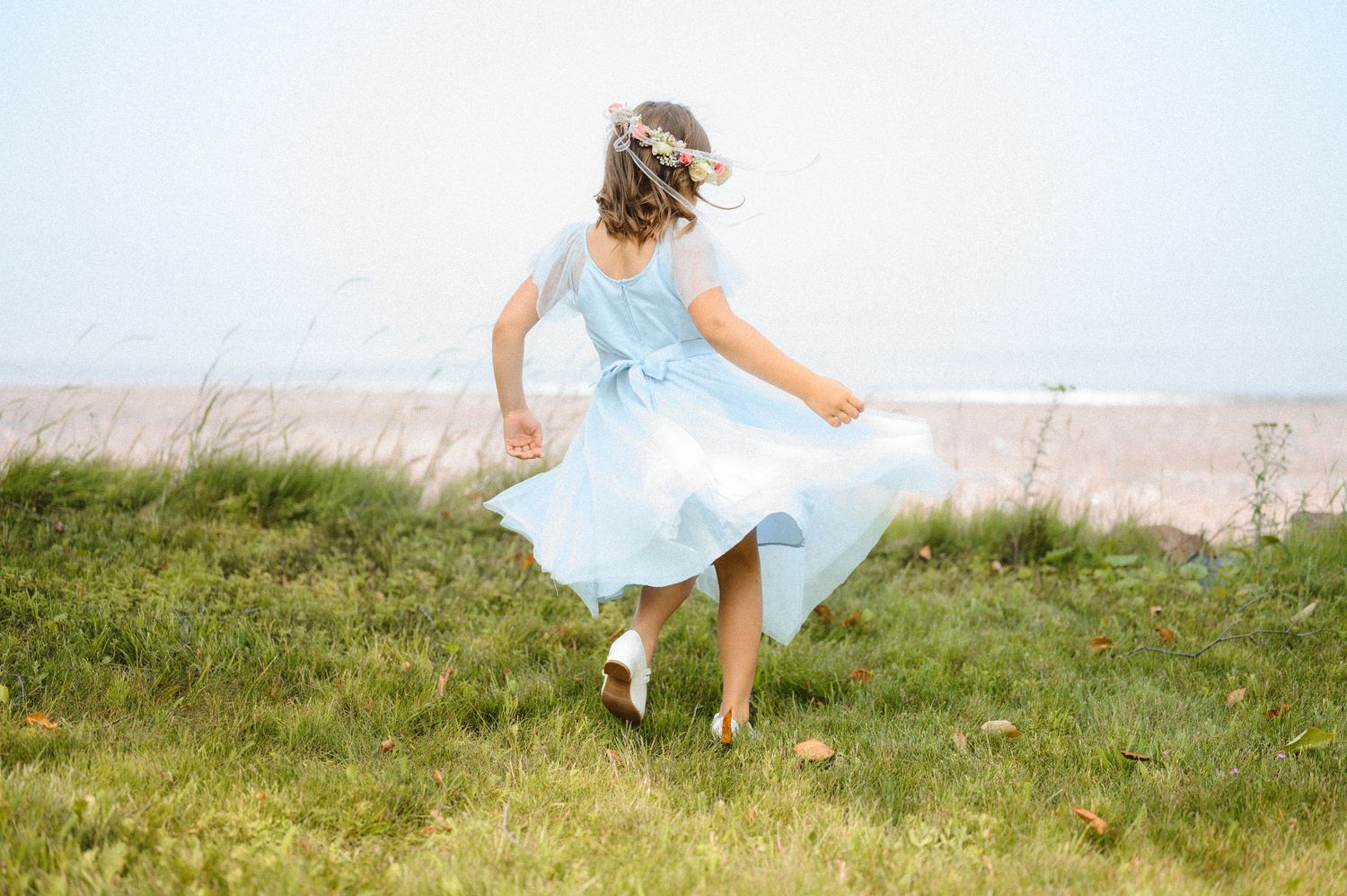 A young girl in a light blue dress and flower crown twirls on a grassy hill near the beach.