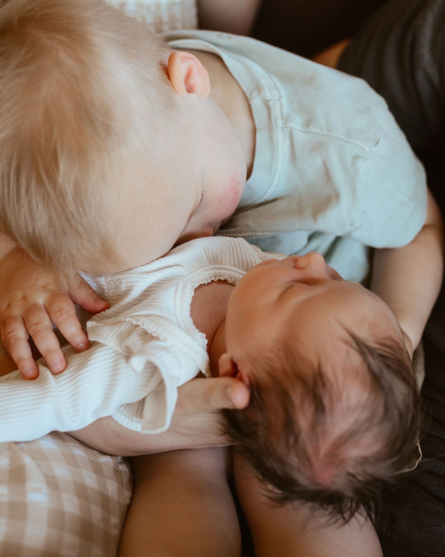 Two babies cuddling closely together, one blonde toddler gently nuzzling a sleeping dark-haired newborn.
