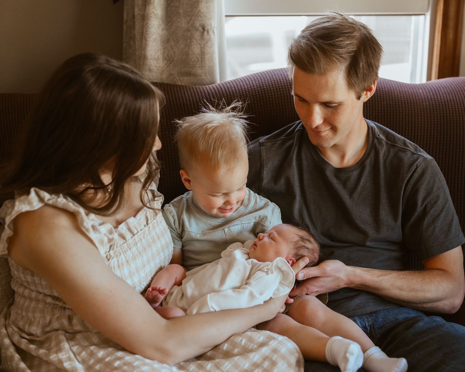 Family of four with newborn baby, toddler, mother and father sitting together on a couch near a window.
