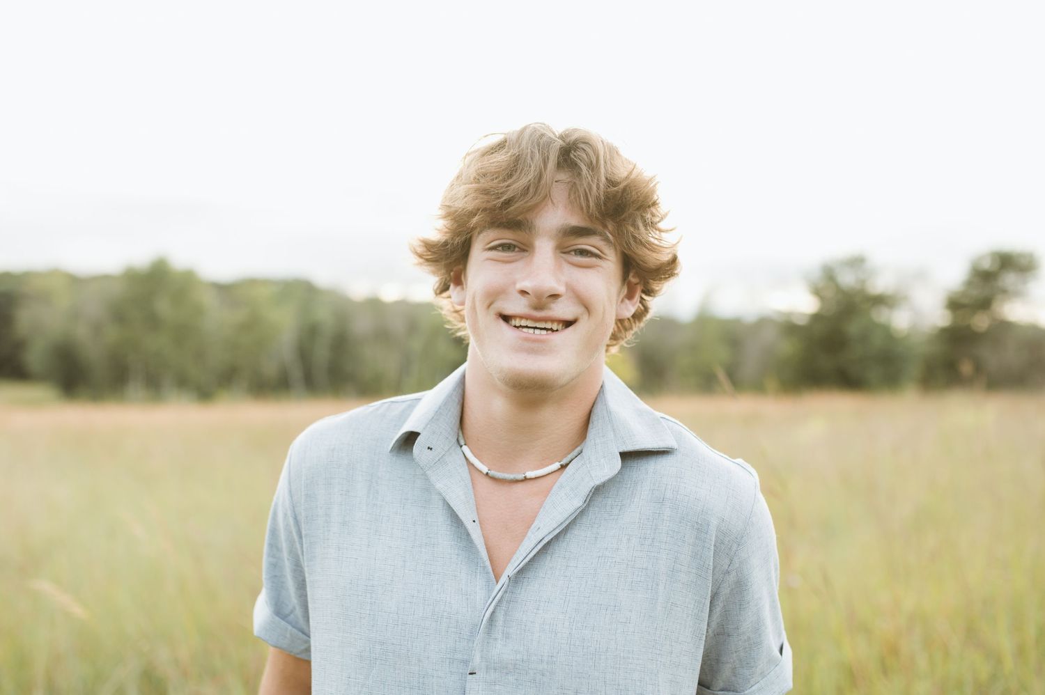 Smiling young man with wavy hair wearing a gray shirt in an open grassy field.