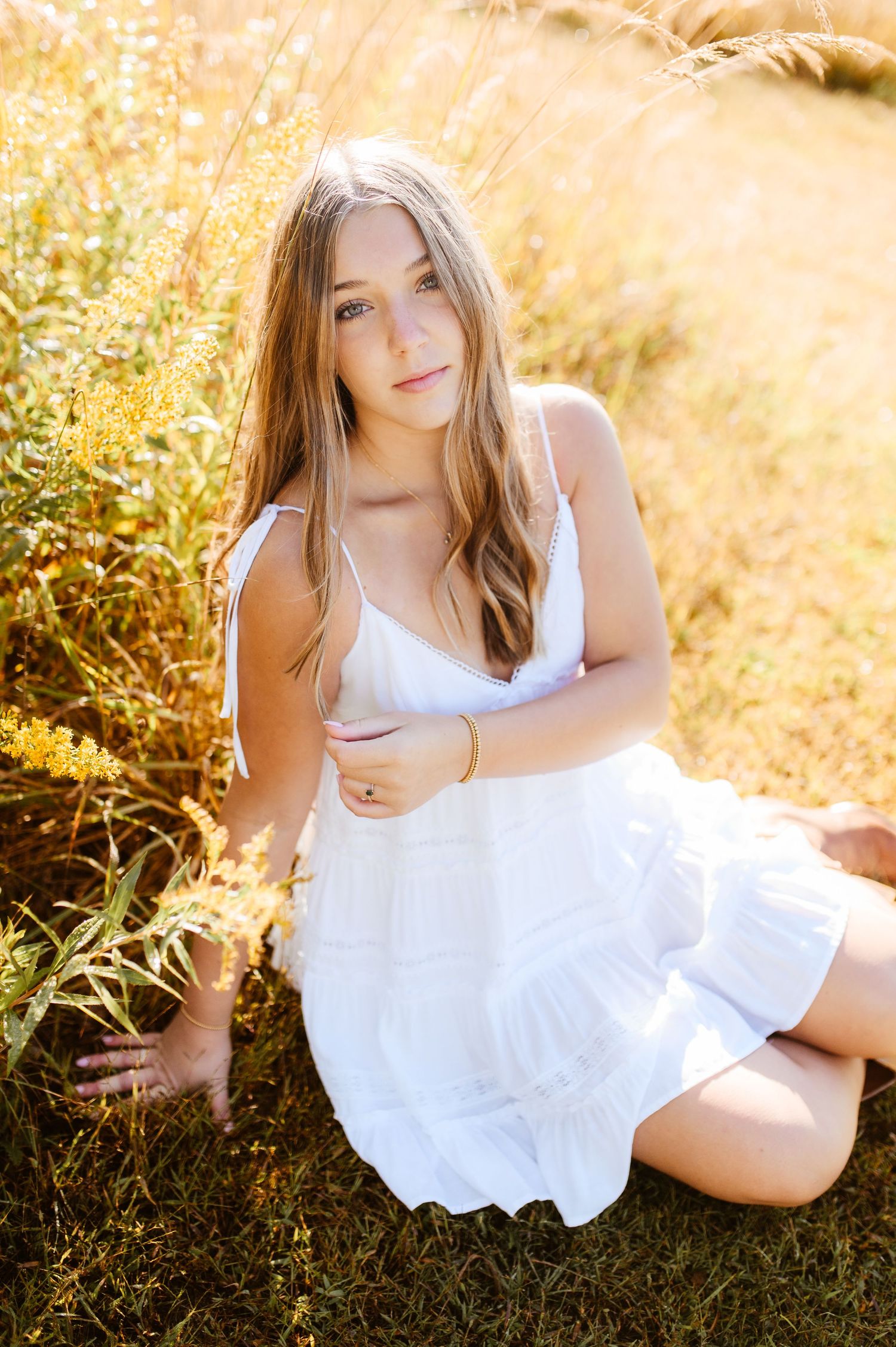 Young woman in white sundress sits gracefully in sunlit golden grass field beside yellow wildflowers.
