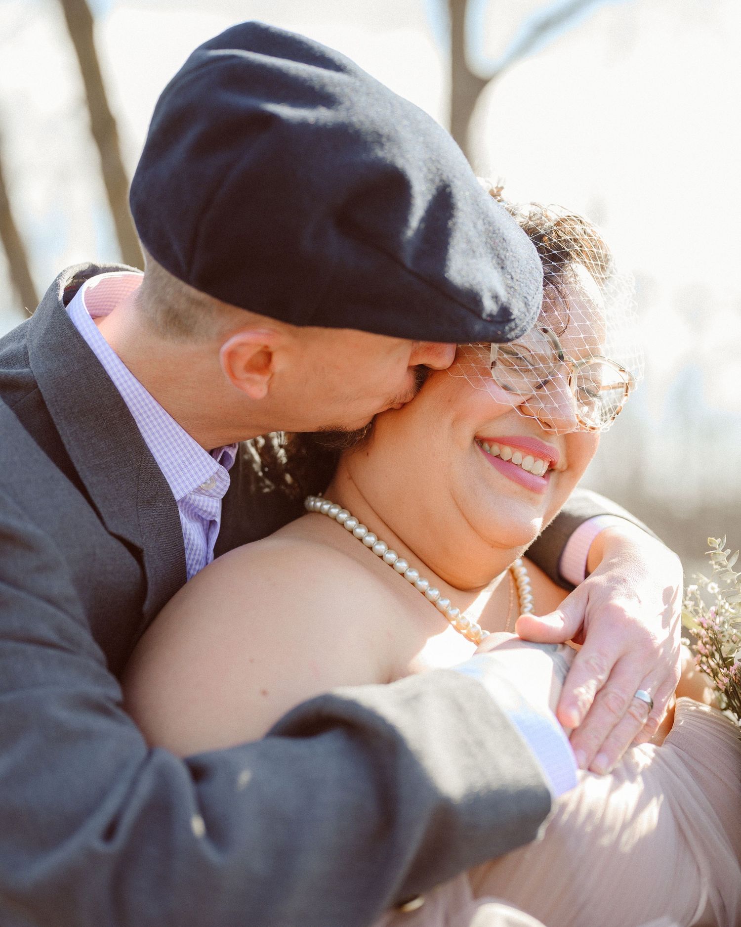 Groom in flat cap kissing smiling bride wearing pearl necklace and birdcage veil outdoors.