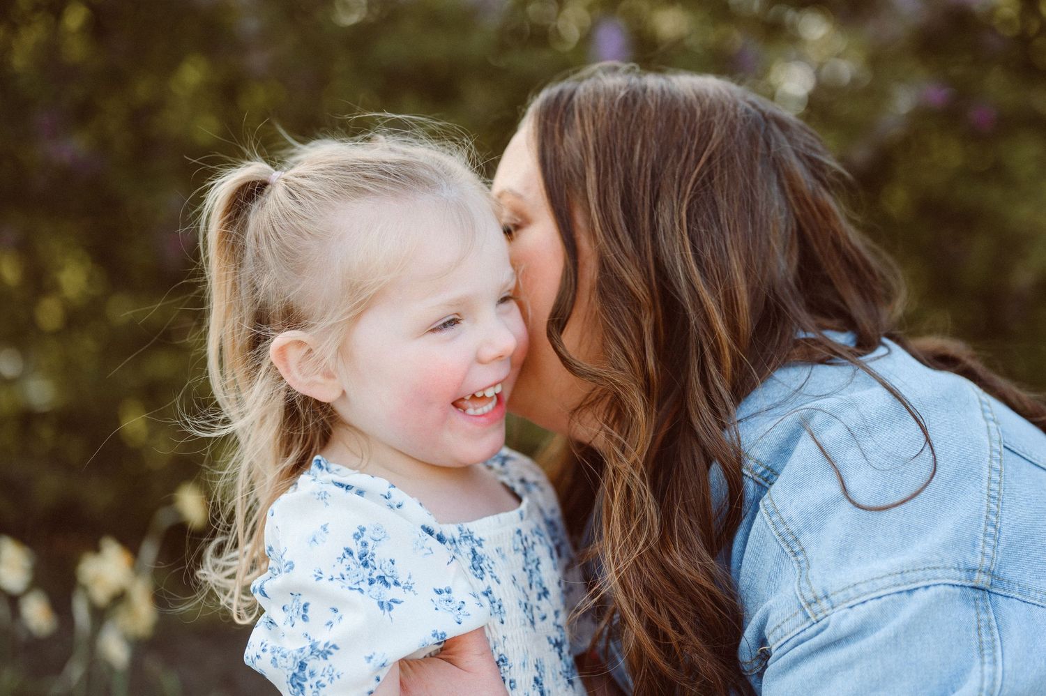 A woman with brown hair kisses the cheek of a smiling blonde toddler girl outdoors.