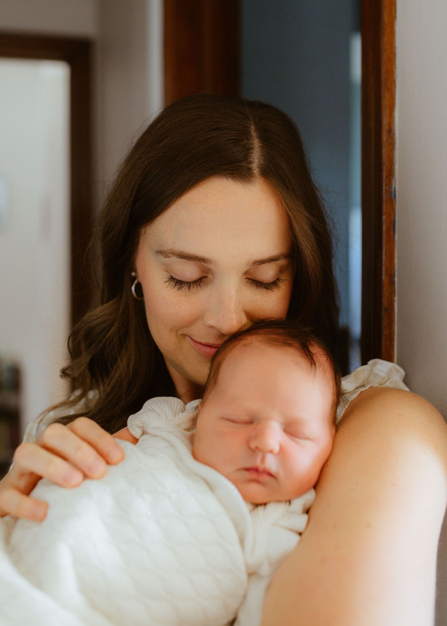 A mother tenderly holds her sleeping newborn baby wrapped in white, smiling softly in a warmly lit room.
