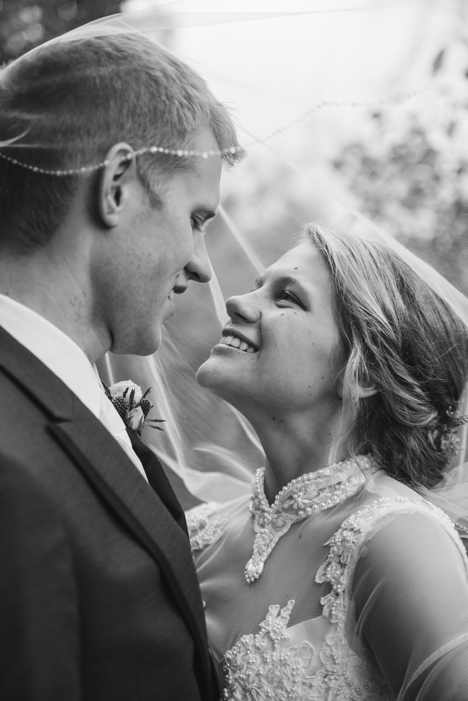 Black and white romantic portrait of smiling bride and groom gazing at each other under bridal veil.