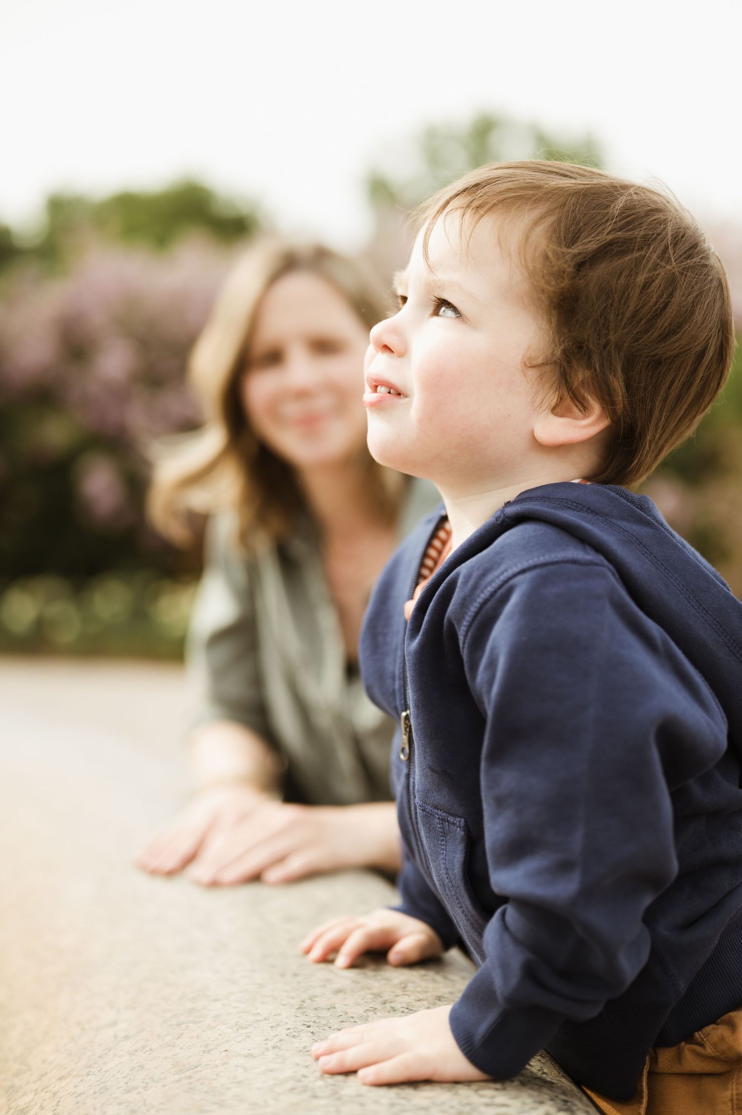 A toddler boy in a navy hoodie looks up curiously while a smiling blonde woman watches him outdoors.